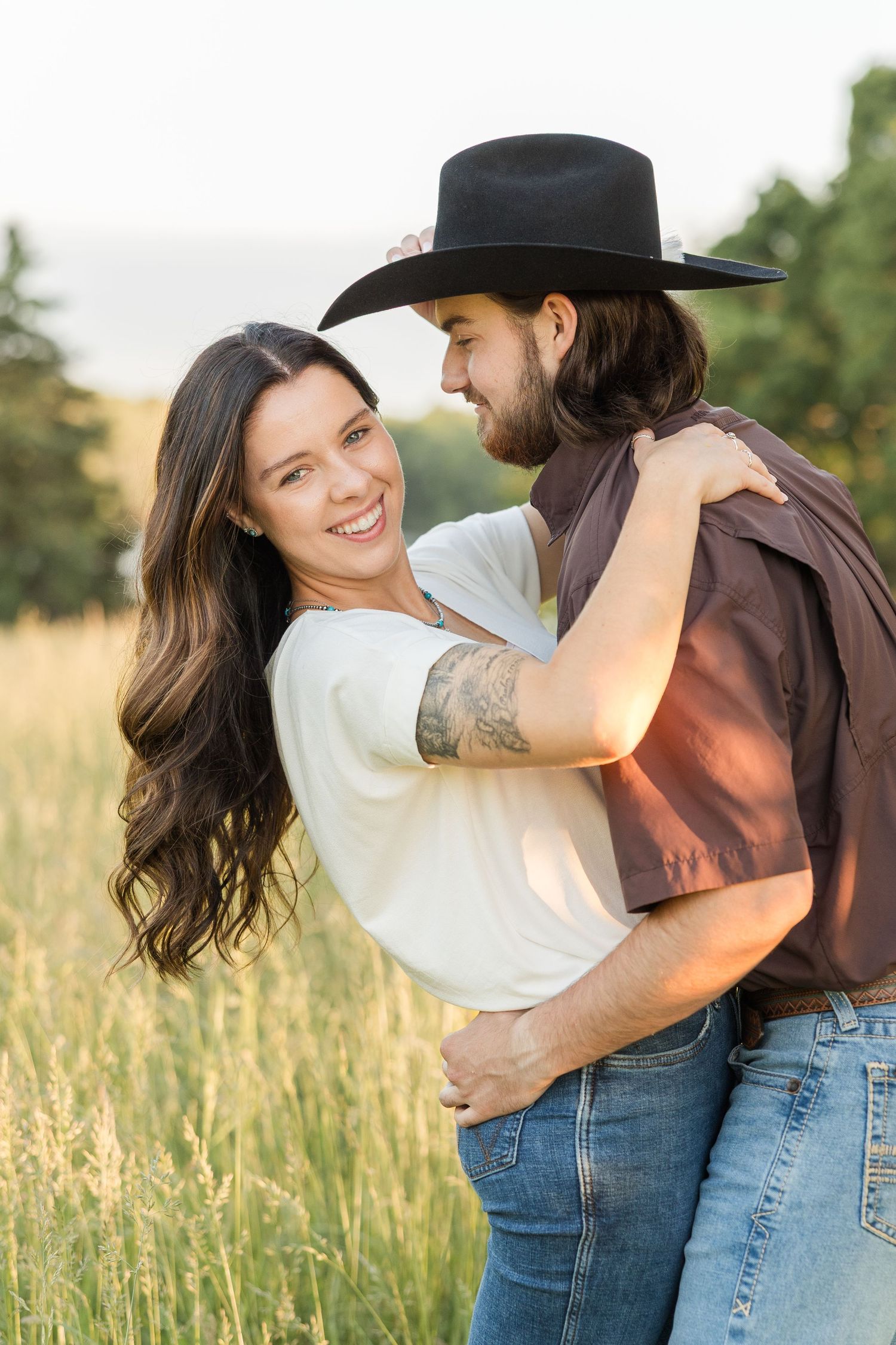 A romantic couple embraces in a sun-lit meadow with tall grass and trees while sharing an intimate moment.