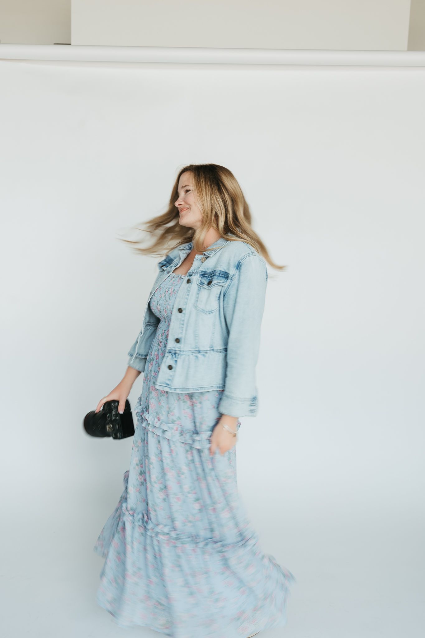 Woman wearing light denim jacket with flowing floral maxi dress twirls joyfully against white wall holding camera.