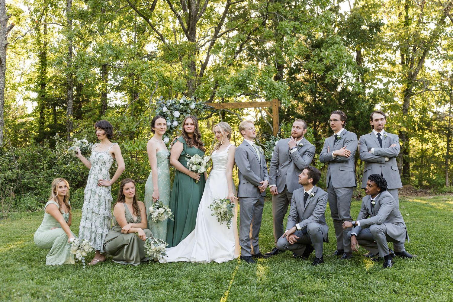 Wedding party poses outdoors in green and gray attire before a floral arch surrounded by lush trees.