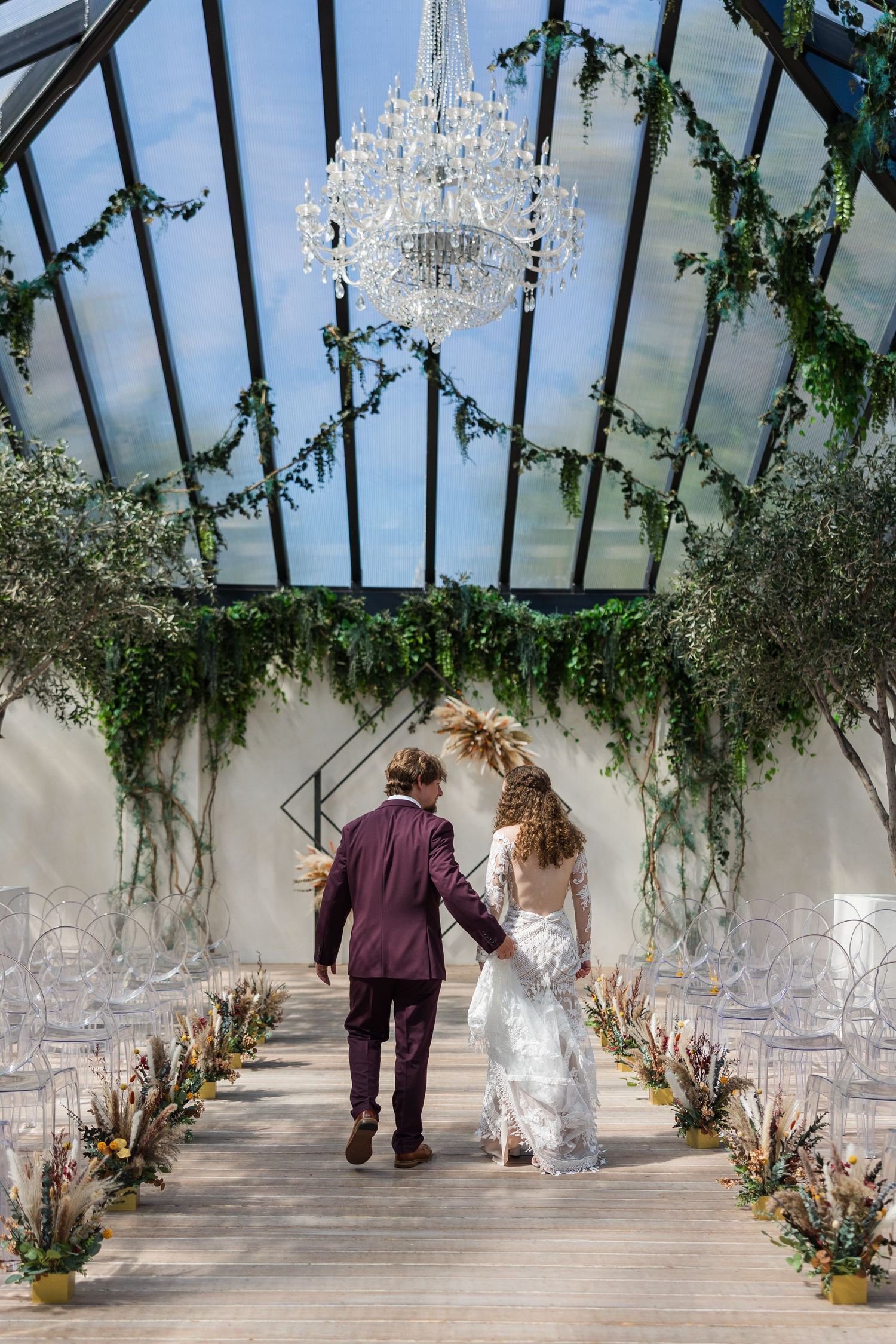 Couple walks hand-in-hand down aisle of glass greenhouse chapel adorned with greenery and chandelier.