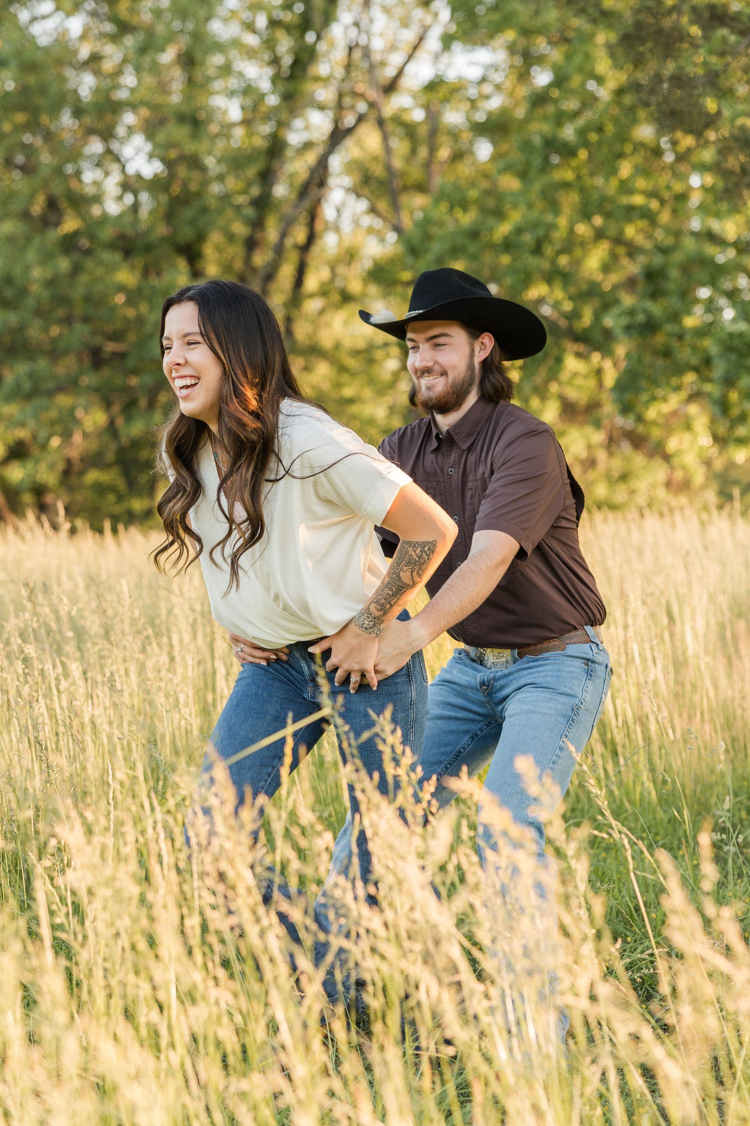 A playful couple runs through a golden field of tall grass during sunset with trees in the background.