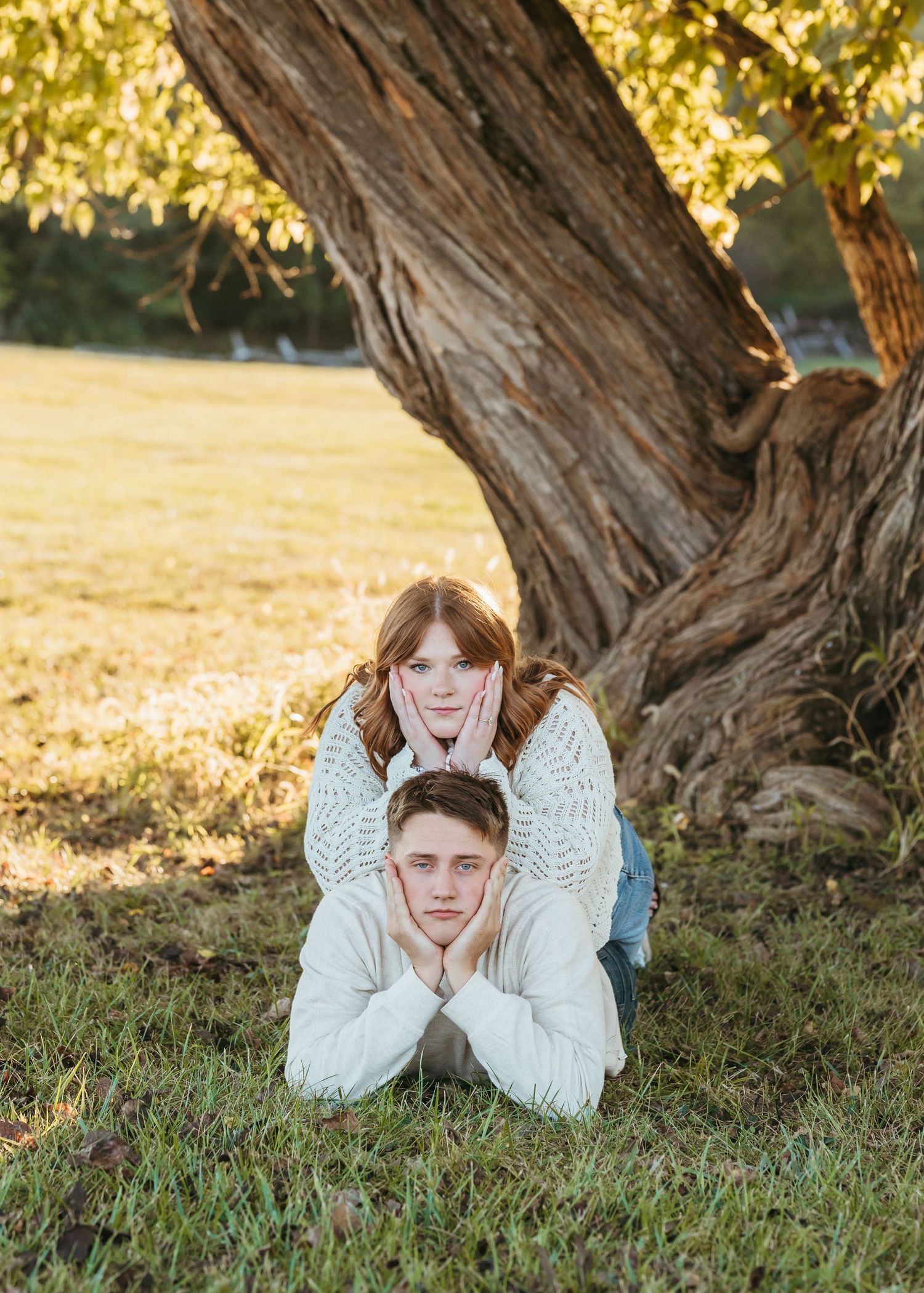 A couple creates silly poses while laying in the grass by a large tree.
