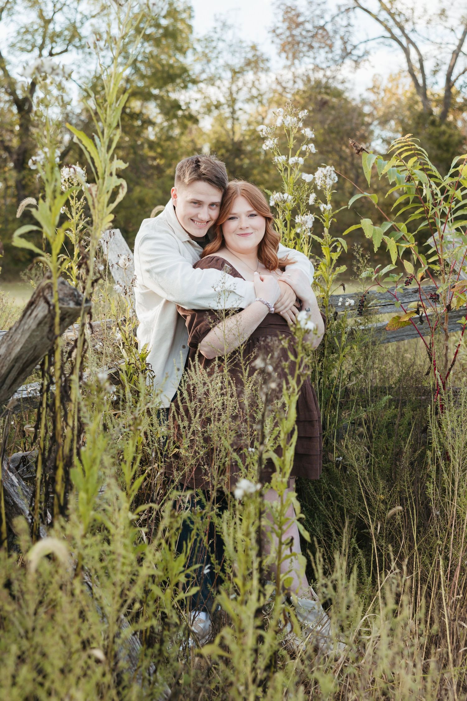 Romantic portrait of a couple embracing by a rustic fence surrounded by tall wildflowers.