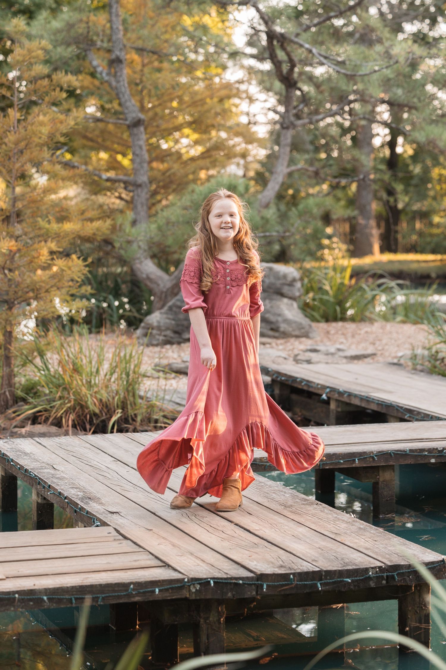 A person in a coral dress twirls on a wooden dock surrounded by autumn trees.