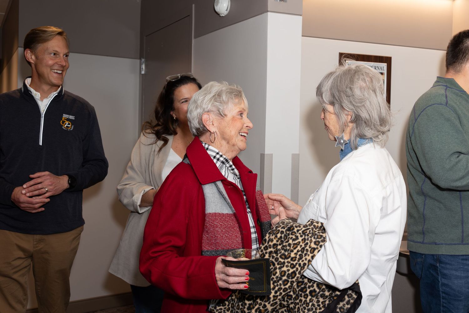 Group socializing in office hallway with person wearing red jacket and plaid scarf.