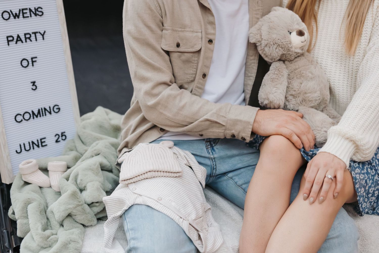 Couple sitting with teddy bear and party announcement sign showing upcoming event details.