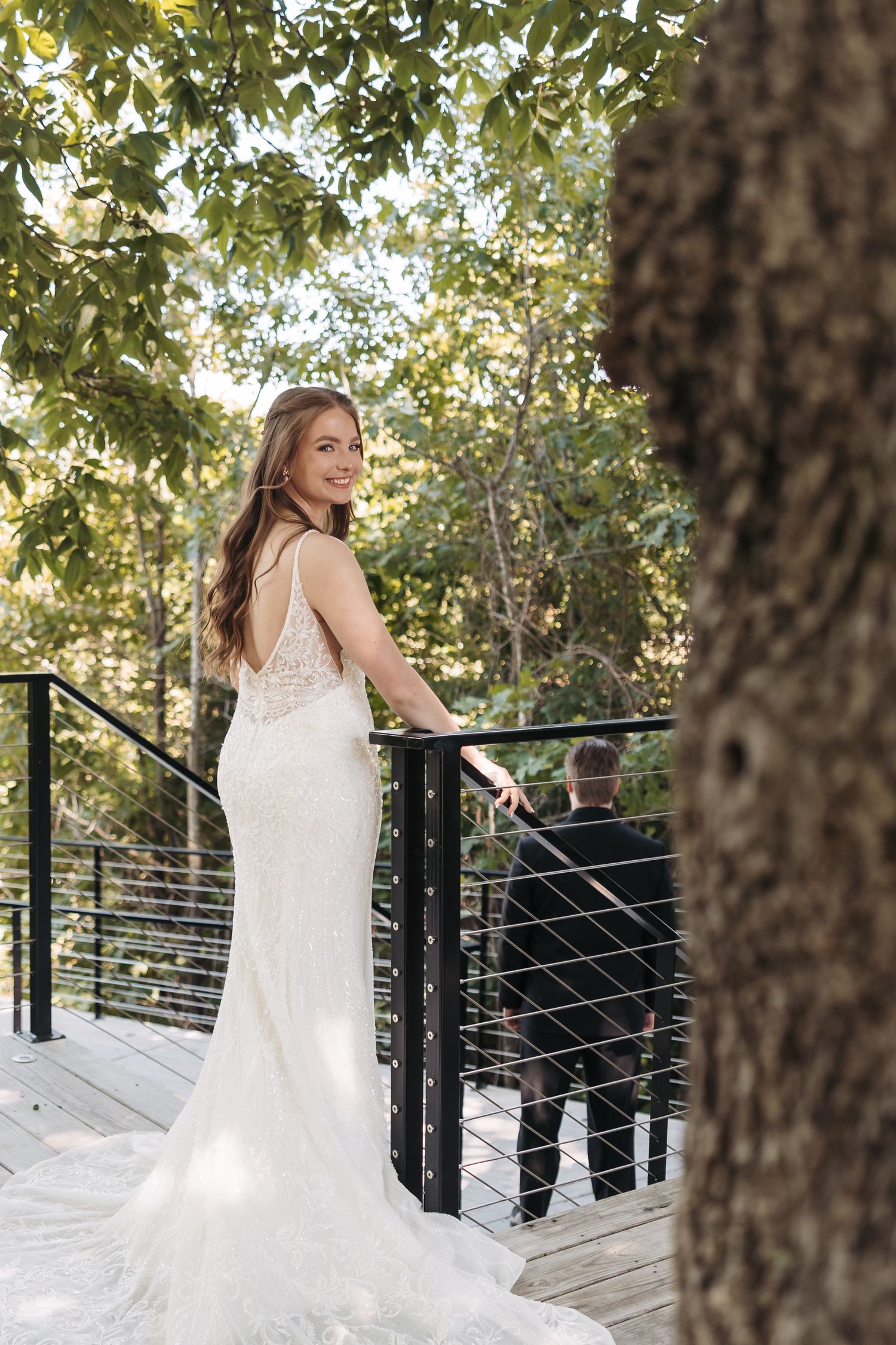 Bride in elegant lace gown poses on wooden deck with flowing train against green foliage.