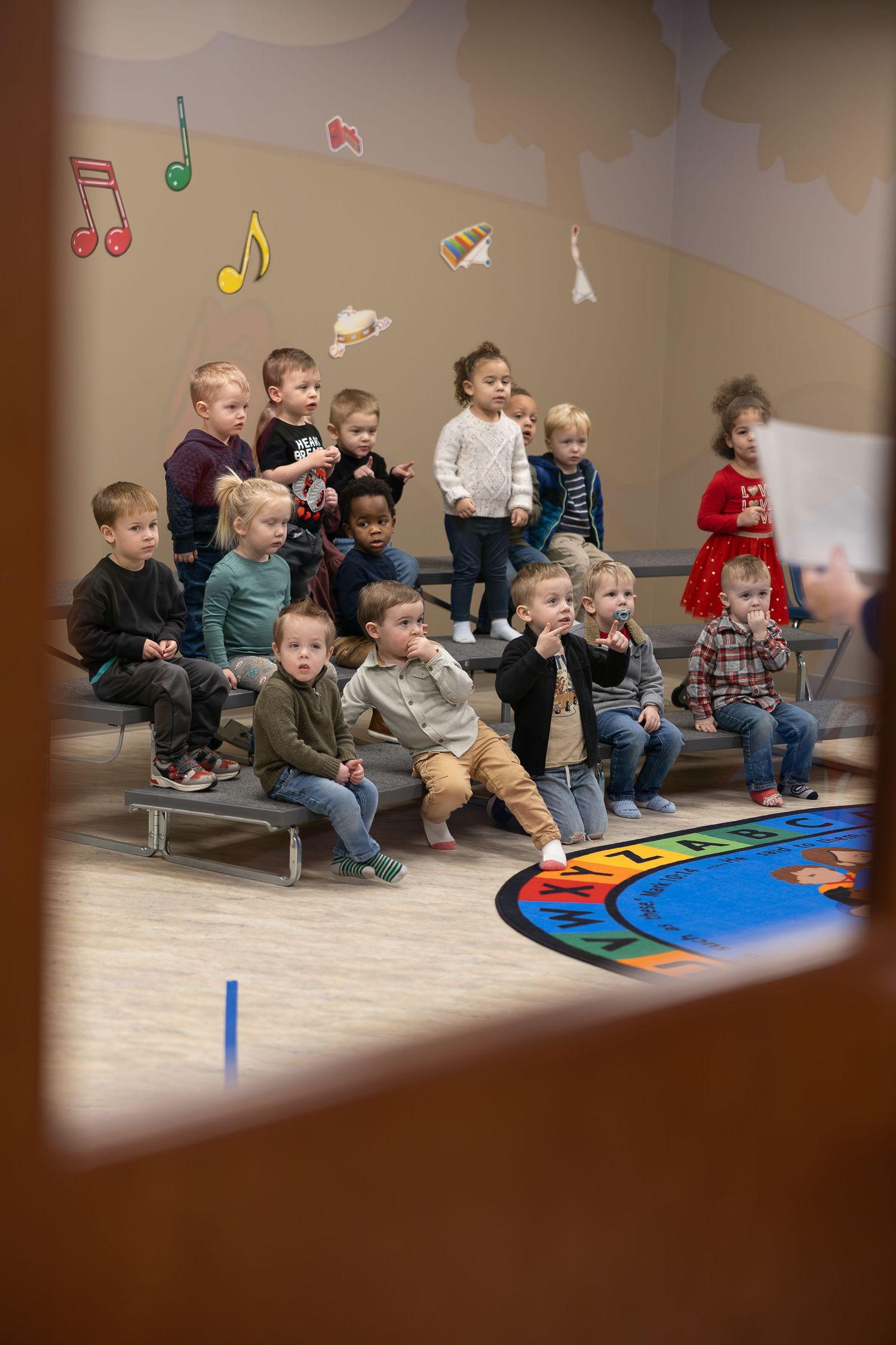 Group of toddlers seated on risers with musical note decorations above.
