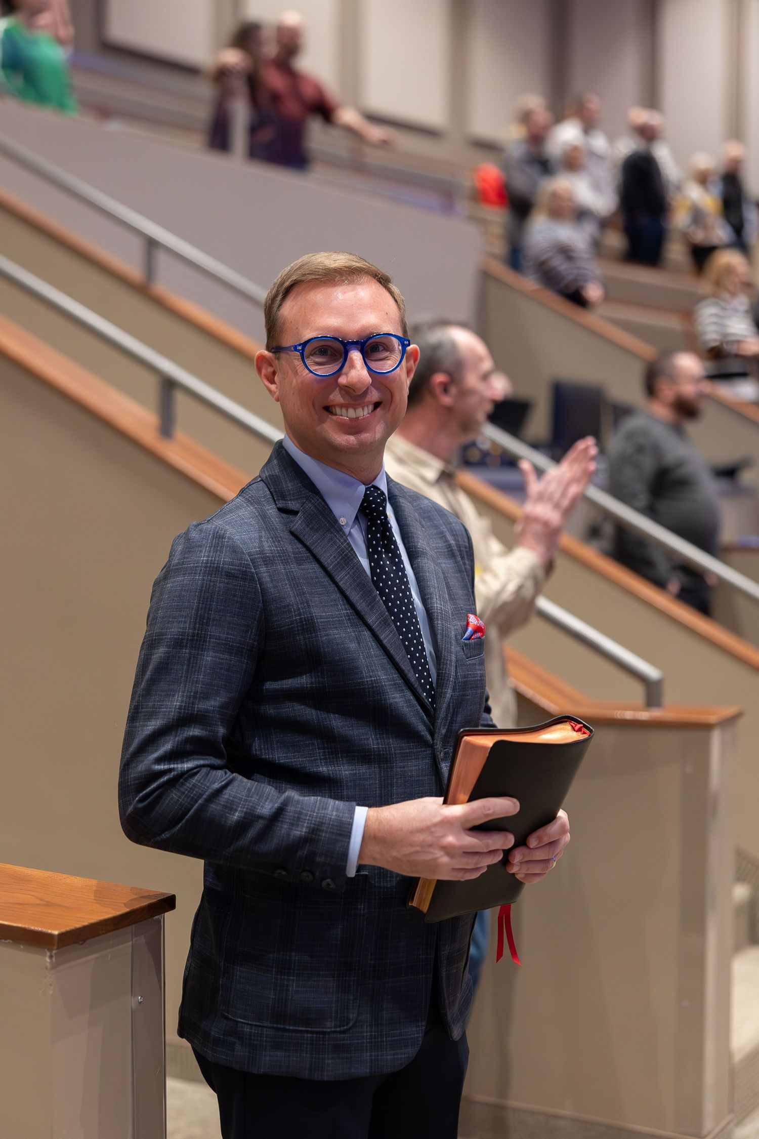 Well-dressed man in suit and glasses holding a book in church setting.