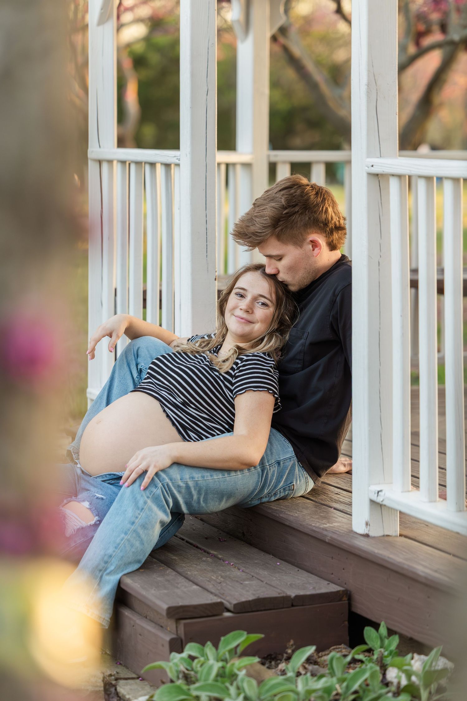 An intimate maternity portrait on a white porch with soft bokeh and garden flowers in the foreground.