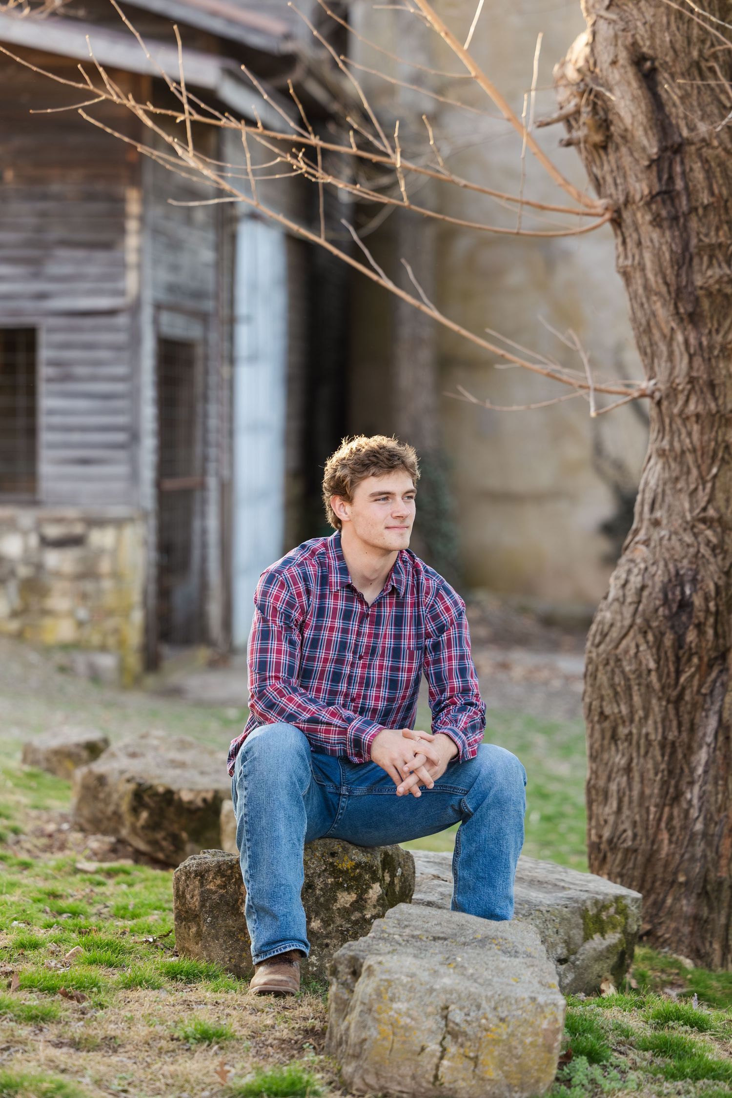 Person in a plaid button-down shirt and jeans sits thoughtfully on a rock near an old rustic building.