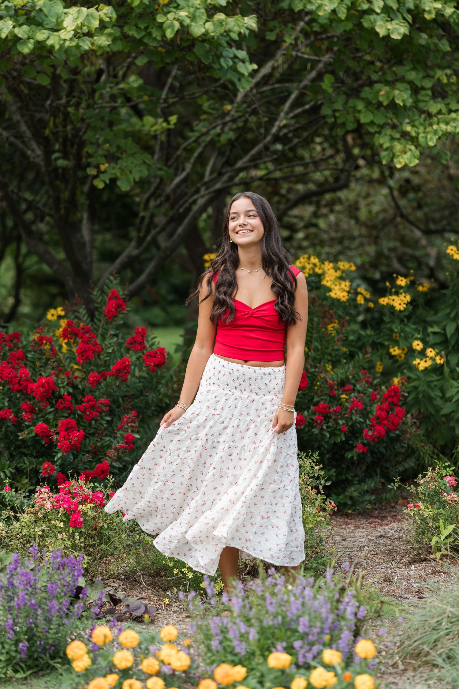 A joyful garden portrait surrounded by red, yellow and purple blooms with a red top and white patterned skirt.