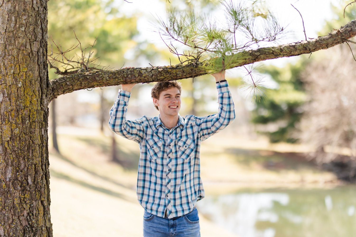 Person in blue plaid shirt holding tree branch near lake on sunny day.