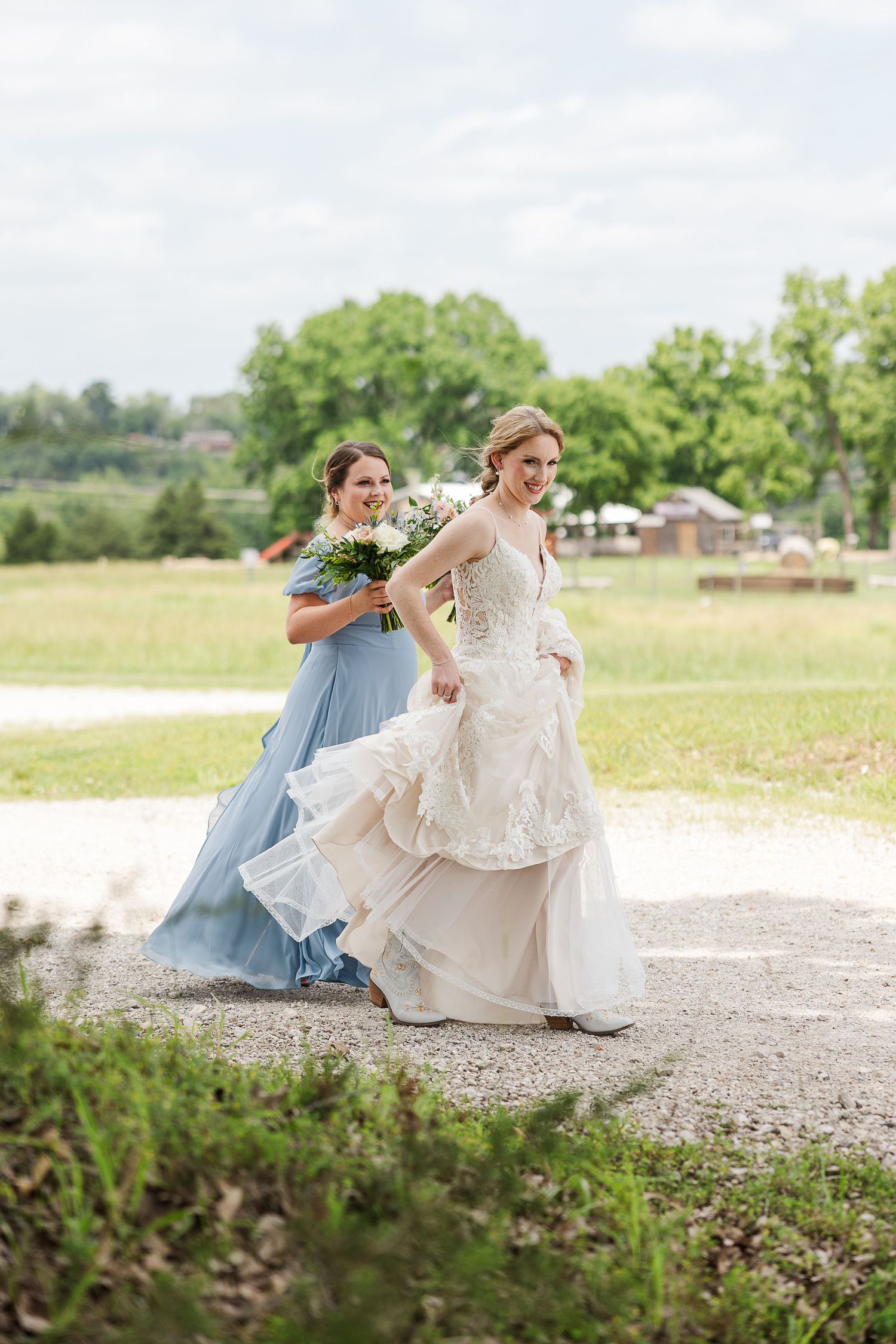A bride in a layered white gown and bridesmaid in light blue dress walk together on a path in a sunny outdoor setting.