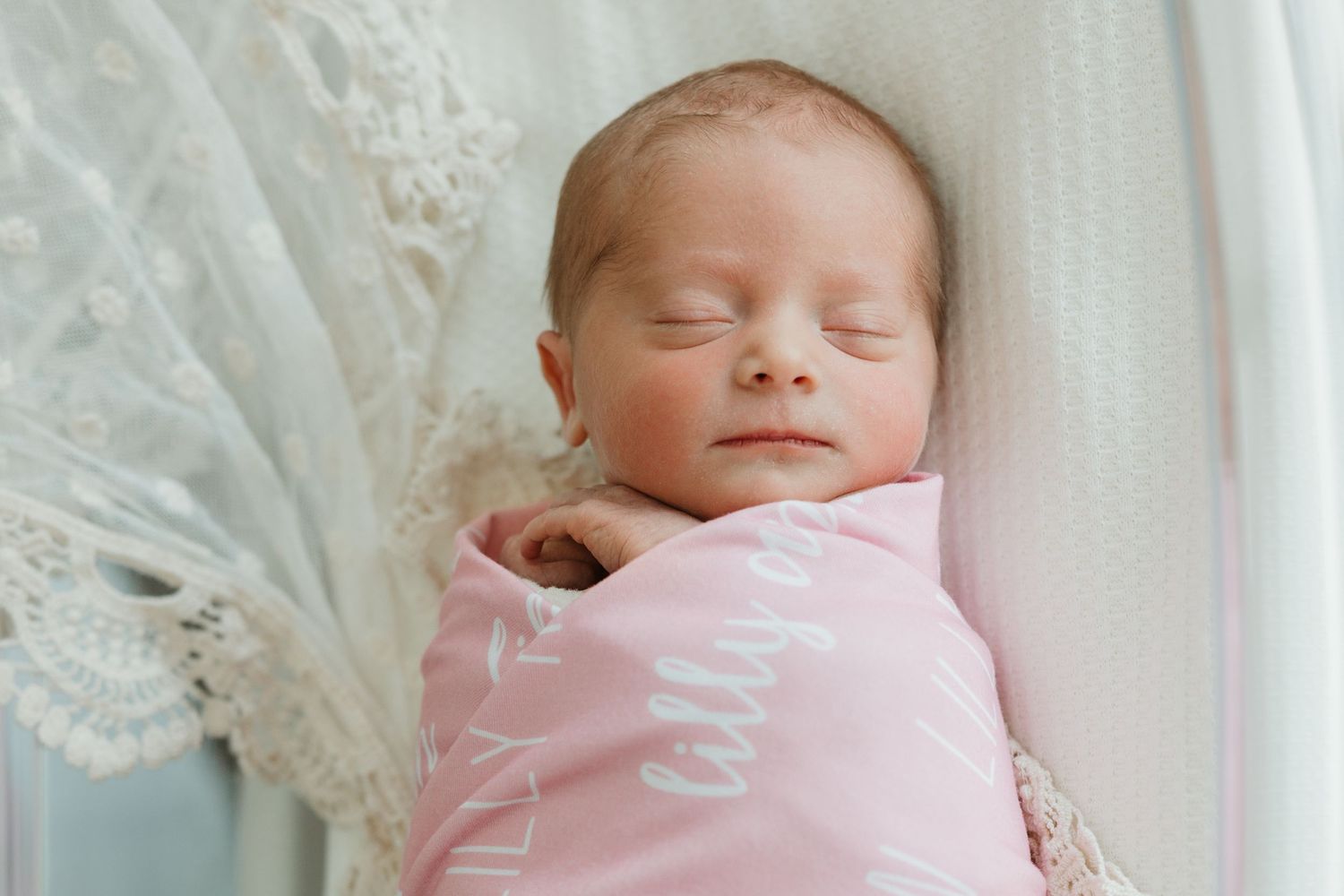 Sleeping newborn baby wrapped in soft pink blanket beside white lace fabric in natural window light.