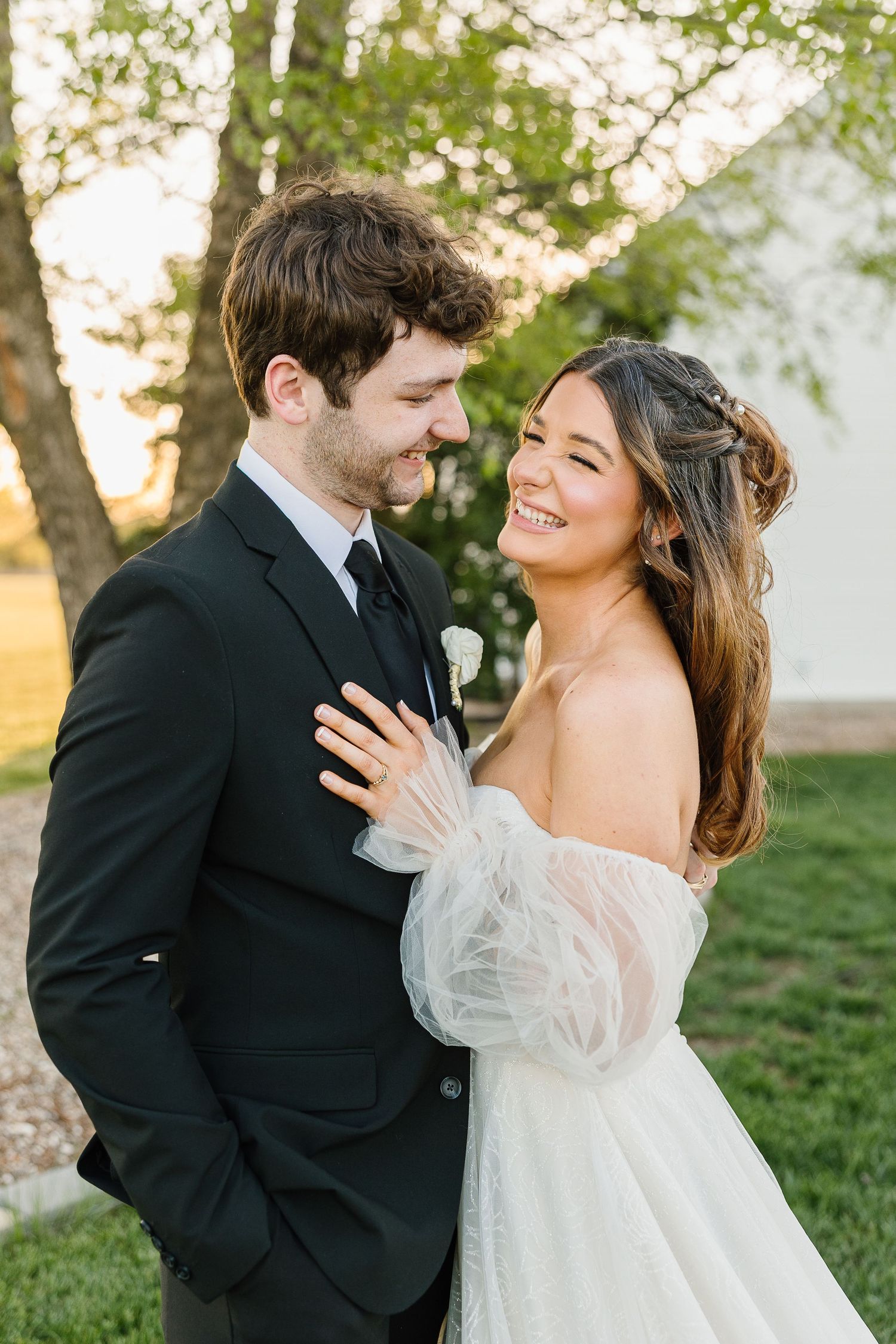 A romantic outdoor wedding photo of a couple sharing a tender moment in front of trees during golden hour.