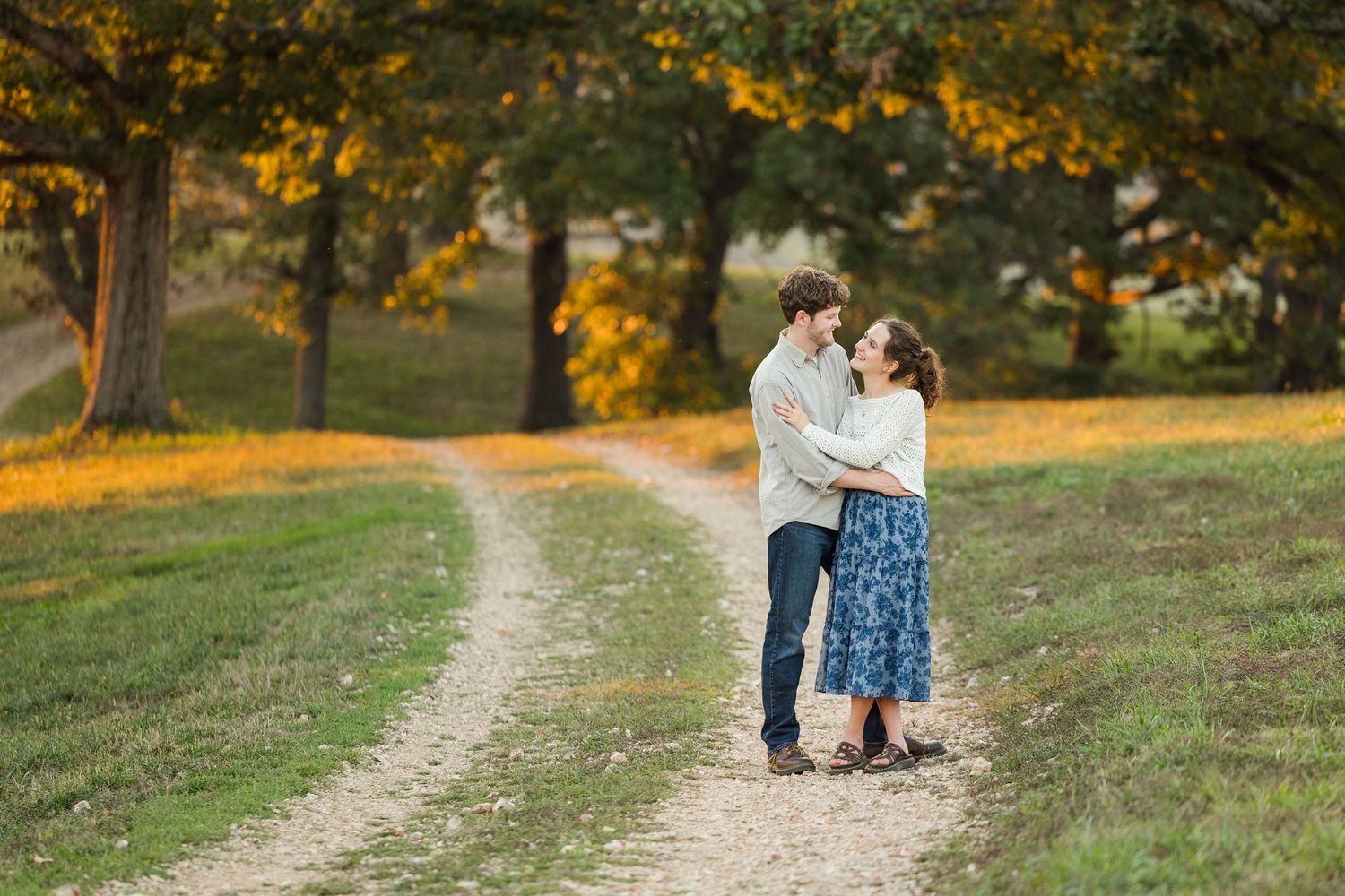 A couple stands together on a winding dirt path during golden hour with autumn trees in the background.