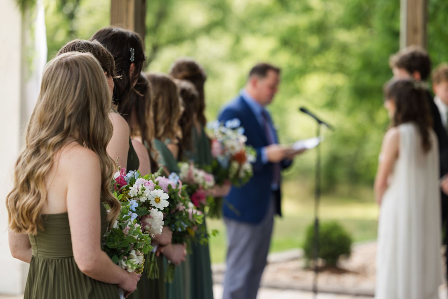 Bridesmaids in green dresses holding floral bouquets stand at an outdoor wedding ceremony with officiant at microphone.