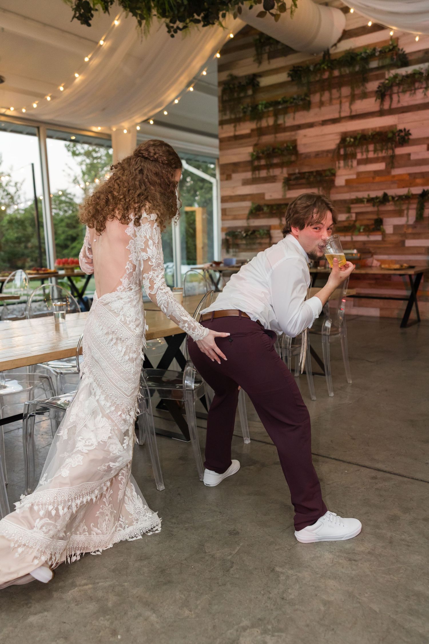 Bride in backless lace gown playfully dances with groom in white shirt at rustic wedding reception.
