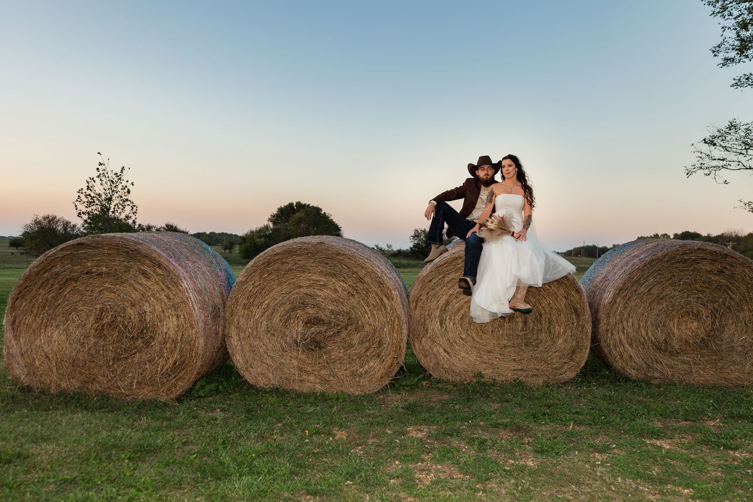 Bride and groom sitting on hay bales at sunset during a rustic country wedding outdoor photo session.