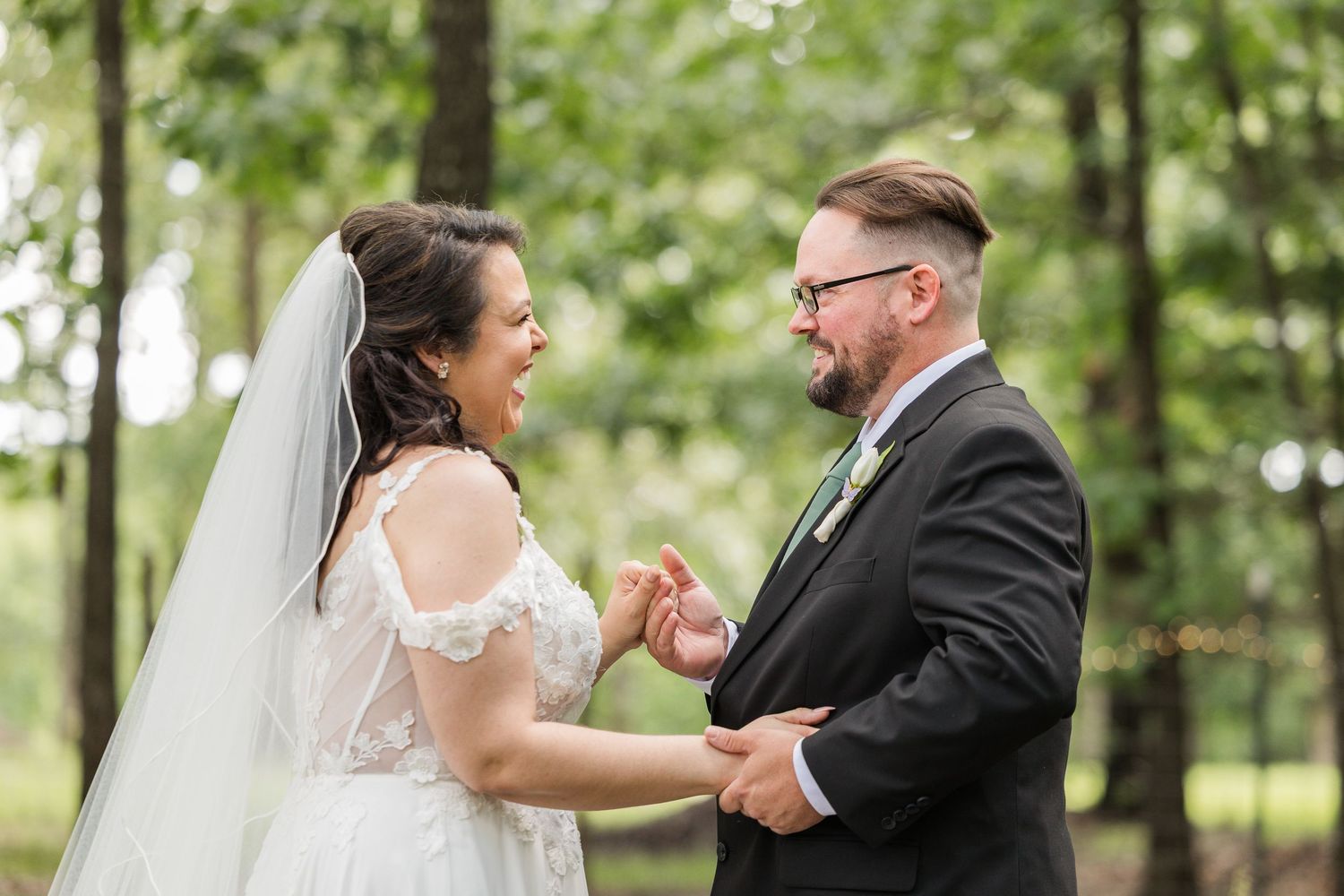 Bride and groom share a joyful moment outdoors, laughing together in a lush green forest setting.