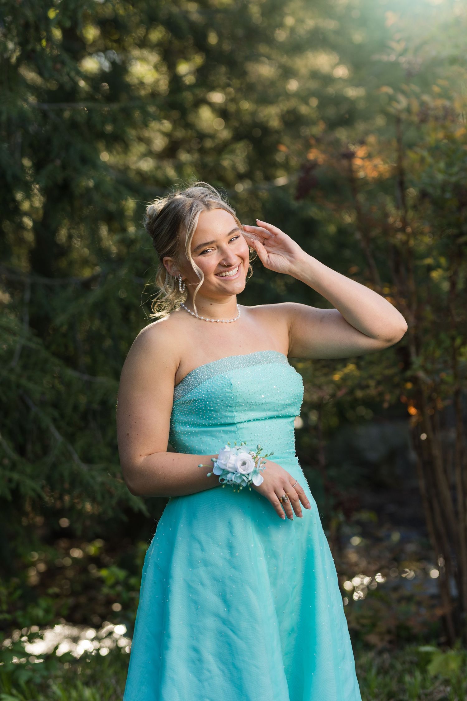 Smiling teenage girl in strapless teal prom dress with wrist corsage and pearl necklace posing outdoors in sunlight.