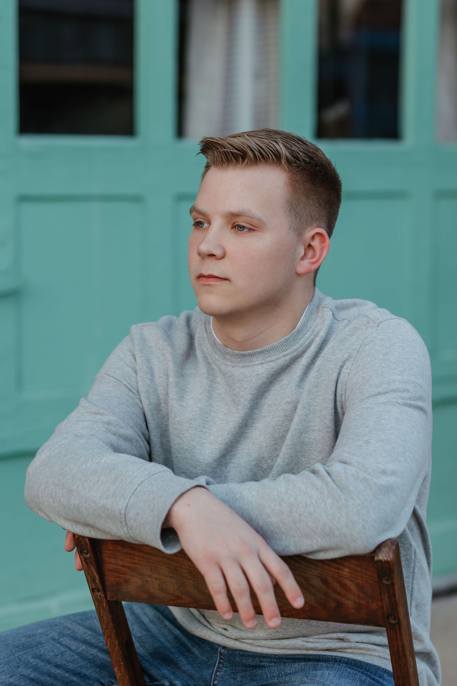 Portrait against turquoise door wearing grey sweatshirt and casual attire.
