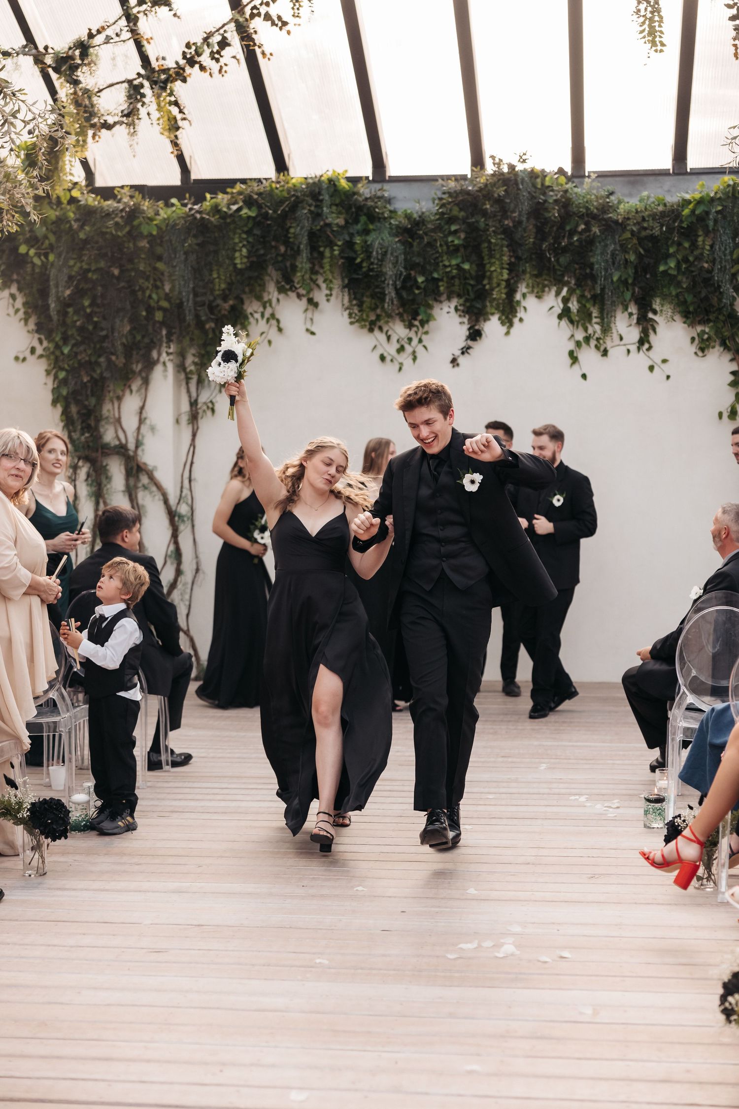 Wedding couple dances joyfully down aisle under greenery-decorated ceiling as guests watch.