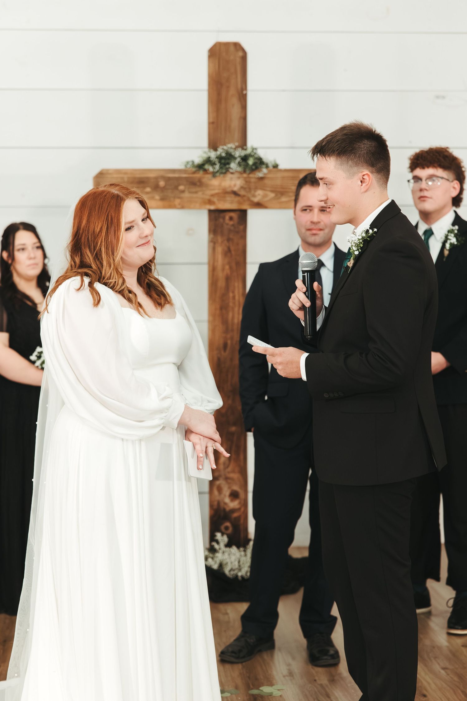 A wedding ceremony takes place in a white chapel with a wooden cross against white shiplap walls.