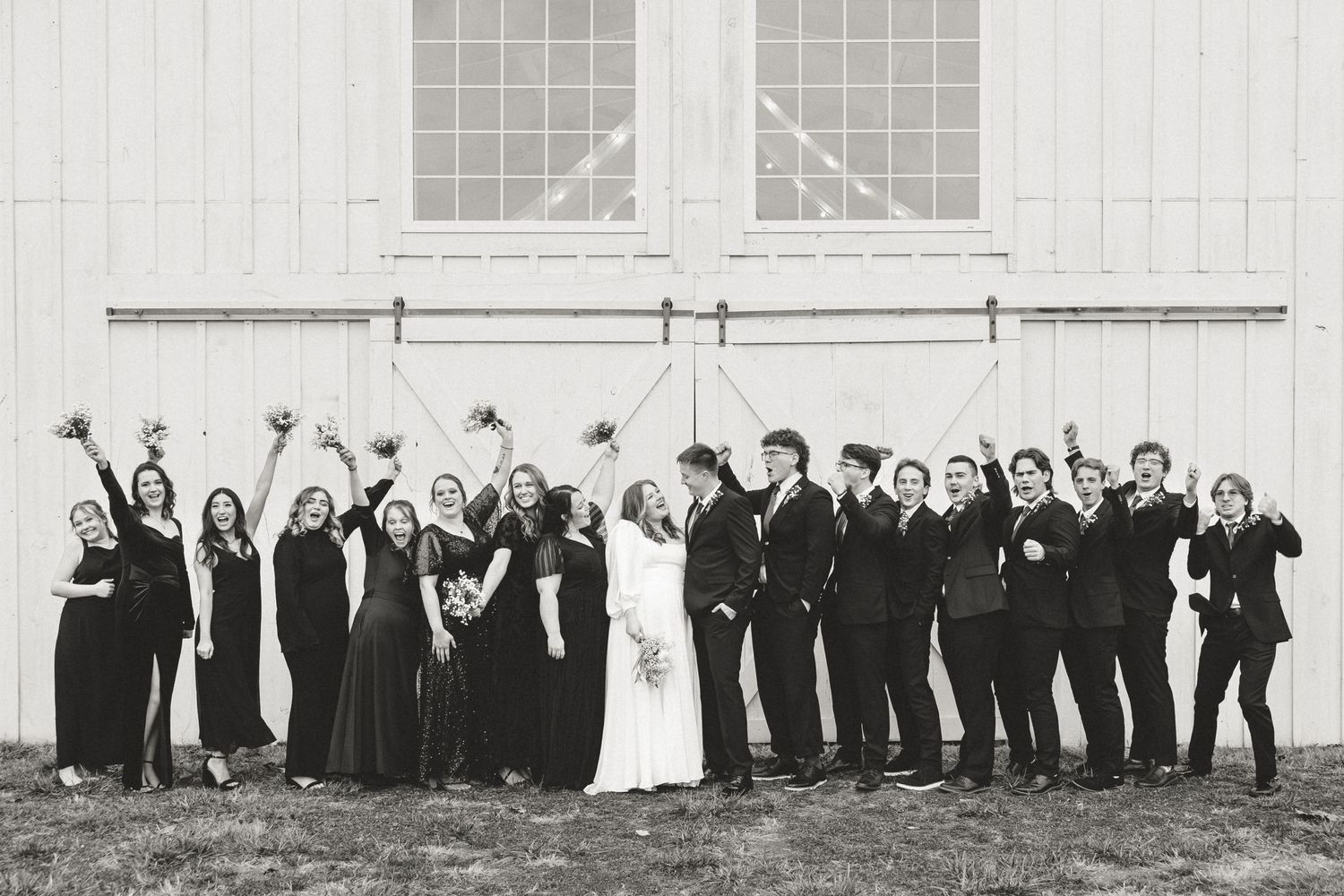 Wedding party poses in a fun lineup against white barn wall, throwing their arms up in celebration.