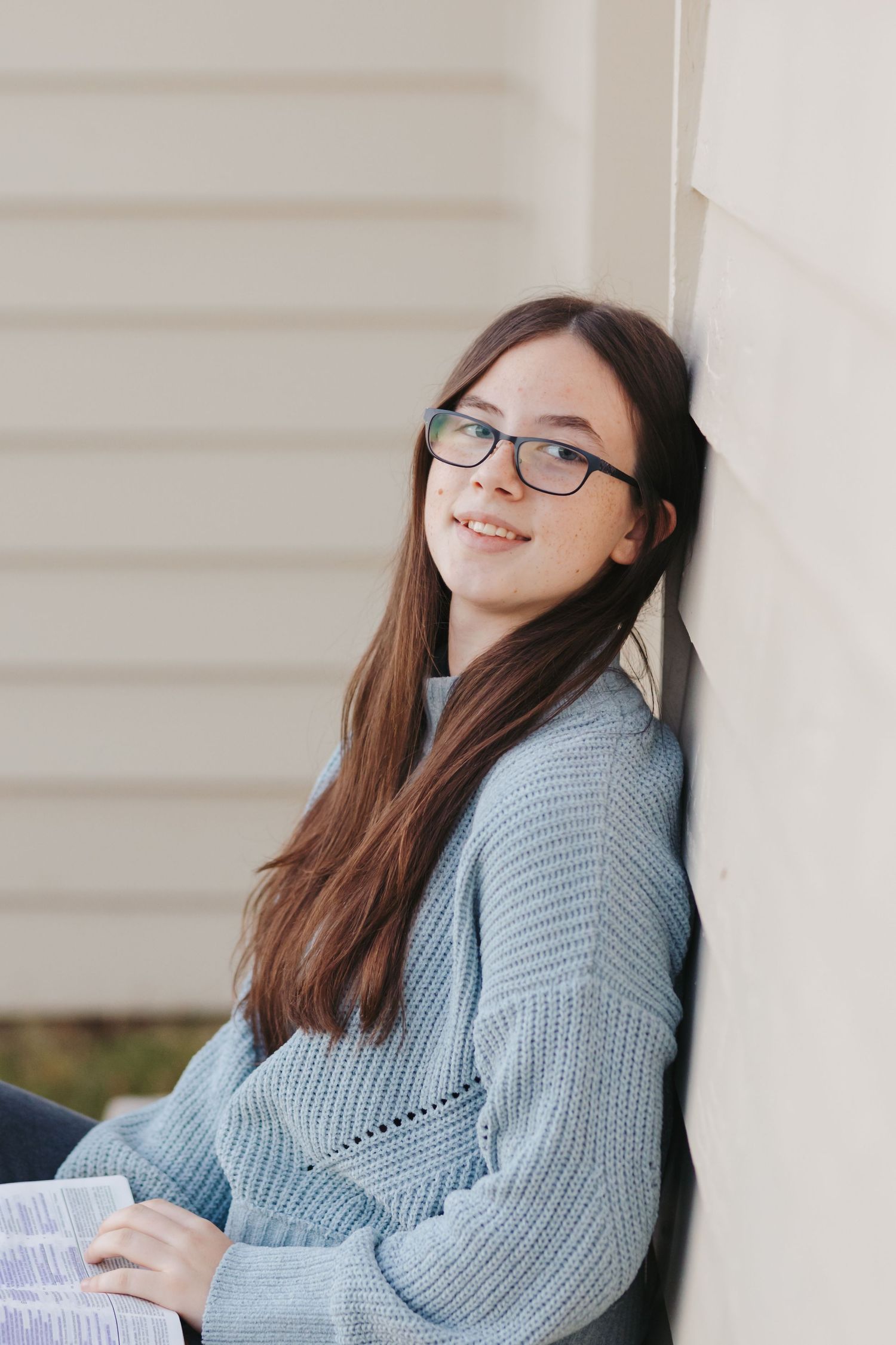 Person in blue sweater and glasses sitting against white wall during photo session.