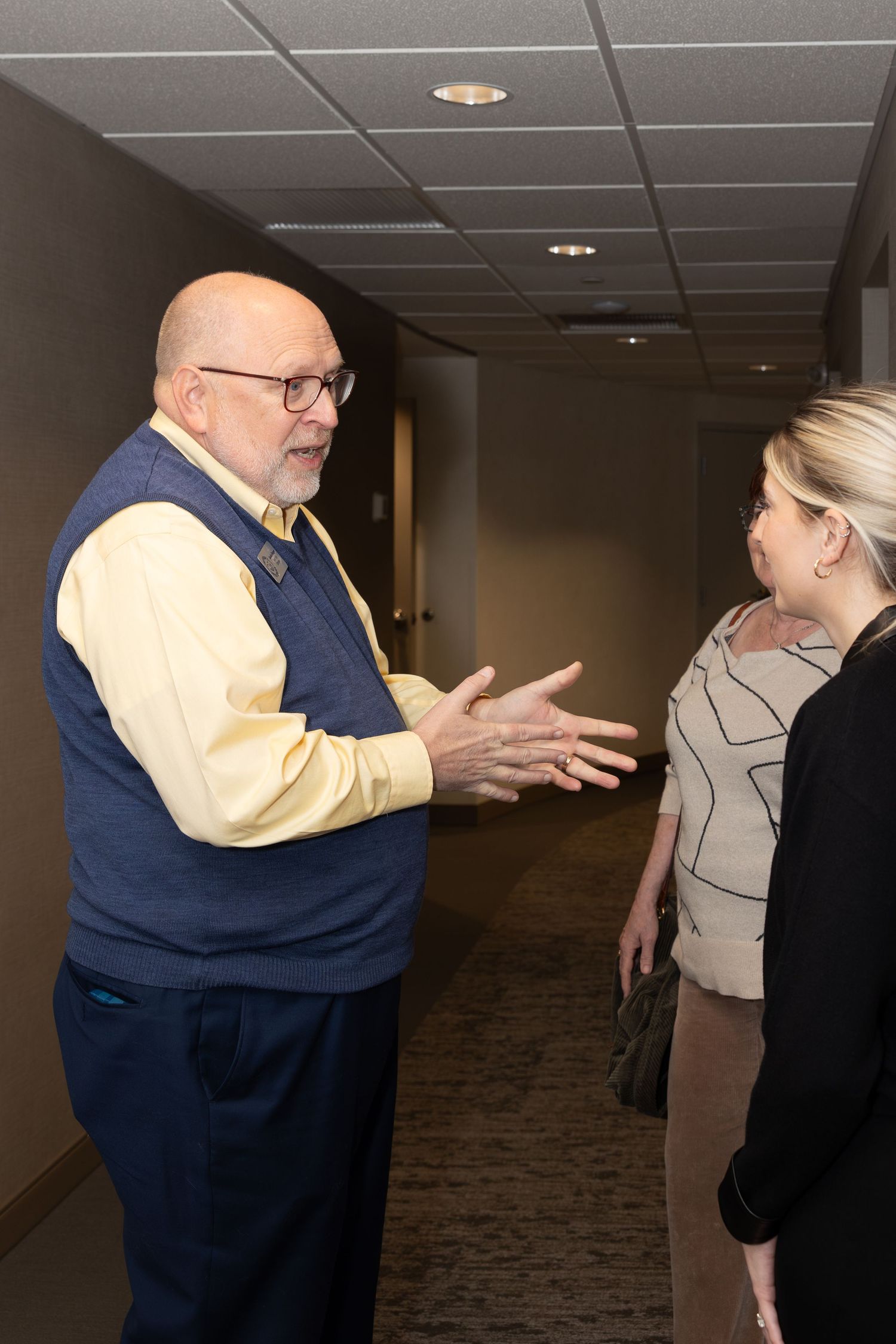 Person in navy sweater vest gesturing while having a conversation in hallway.