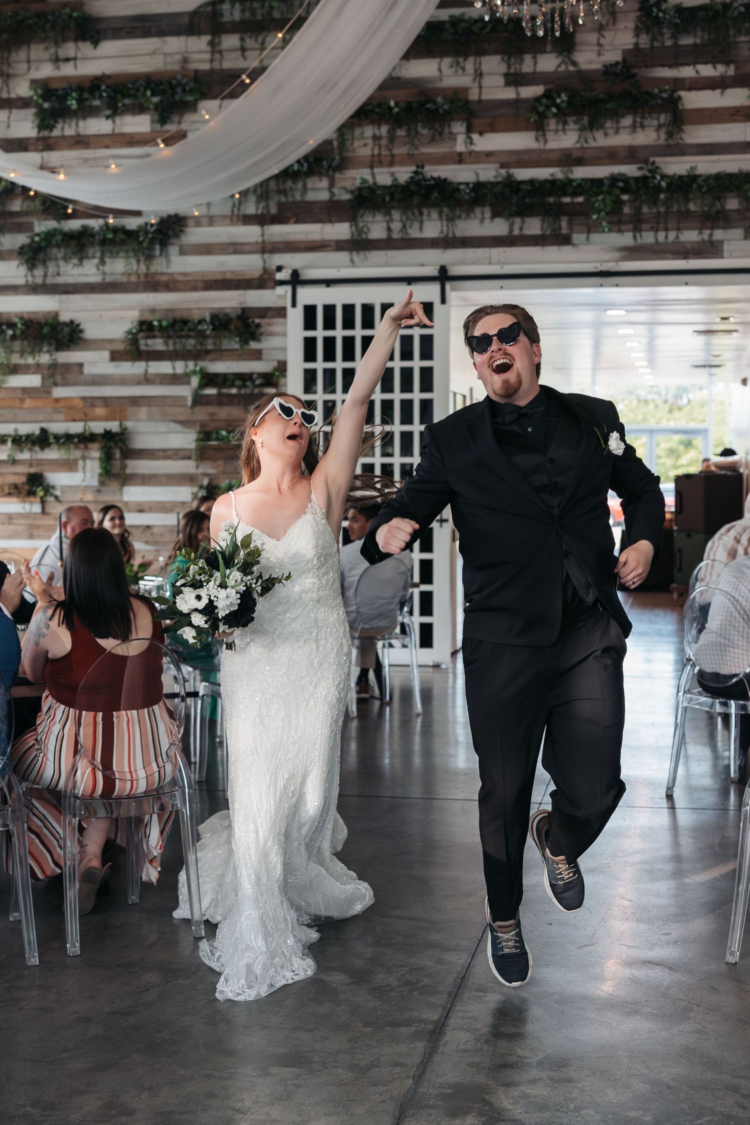 A couple jumps with joy during their wedding reception in an indoor venue with greenery decor.