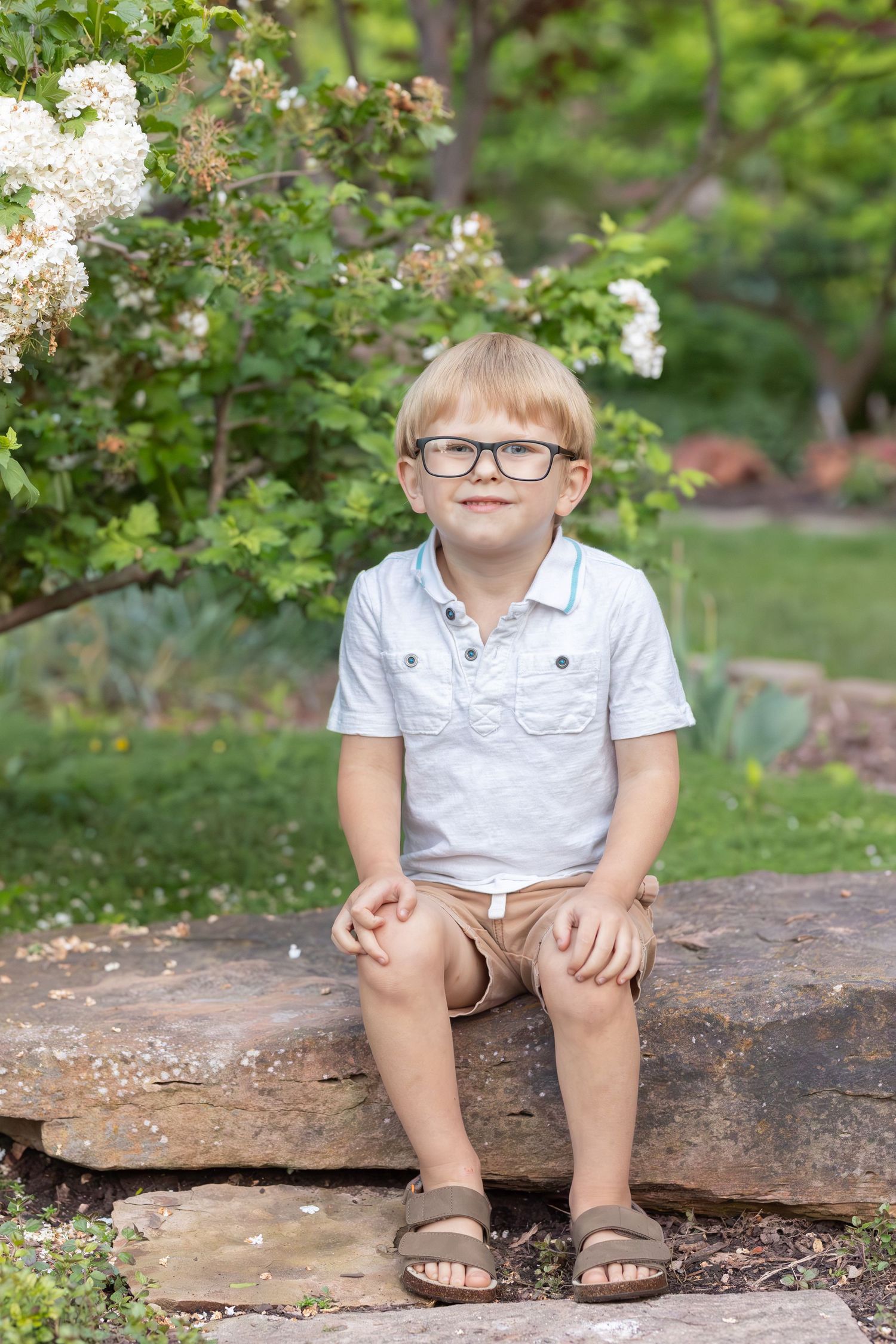 A small child wearing glasses and a white polo shirt sits on a stone ledge in a garden with blooming flowers.