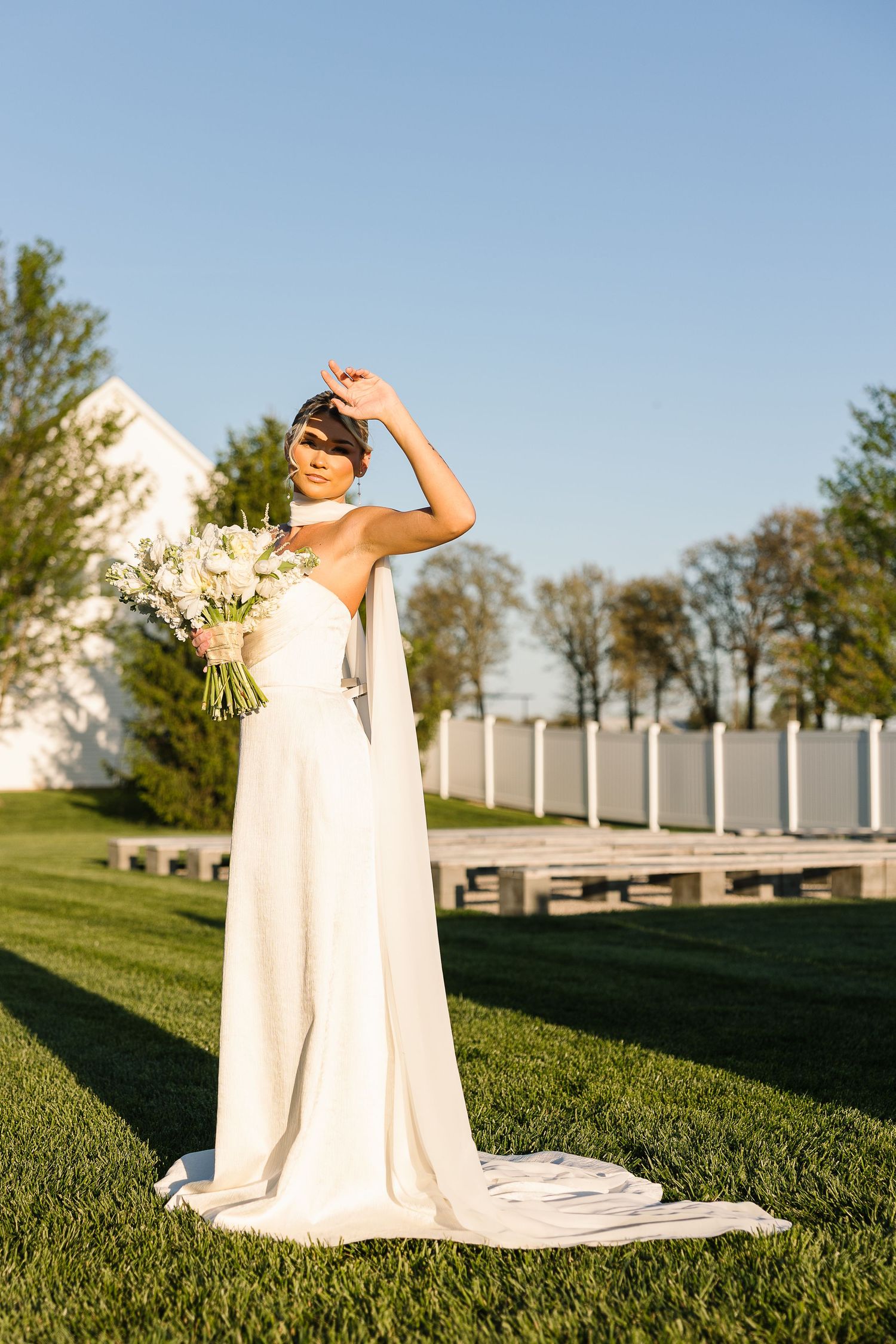 A bride in a flowing white wedding dress stands on a sunny lawn holding a bouquet of flowers against a bright blue sky.