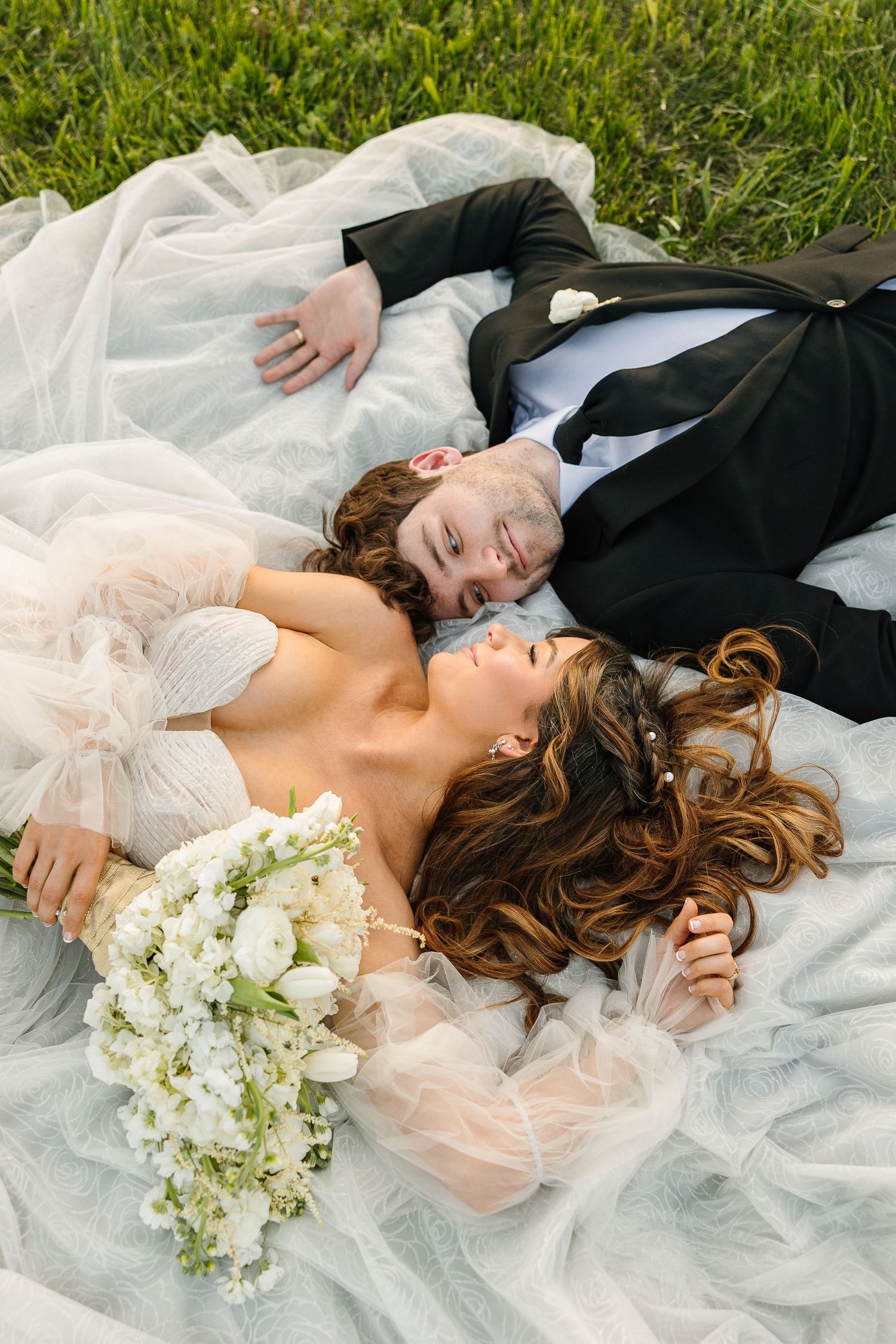 Wedding couple lying together on white tulle with a bouquet of white flowers on grass outdoors.