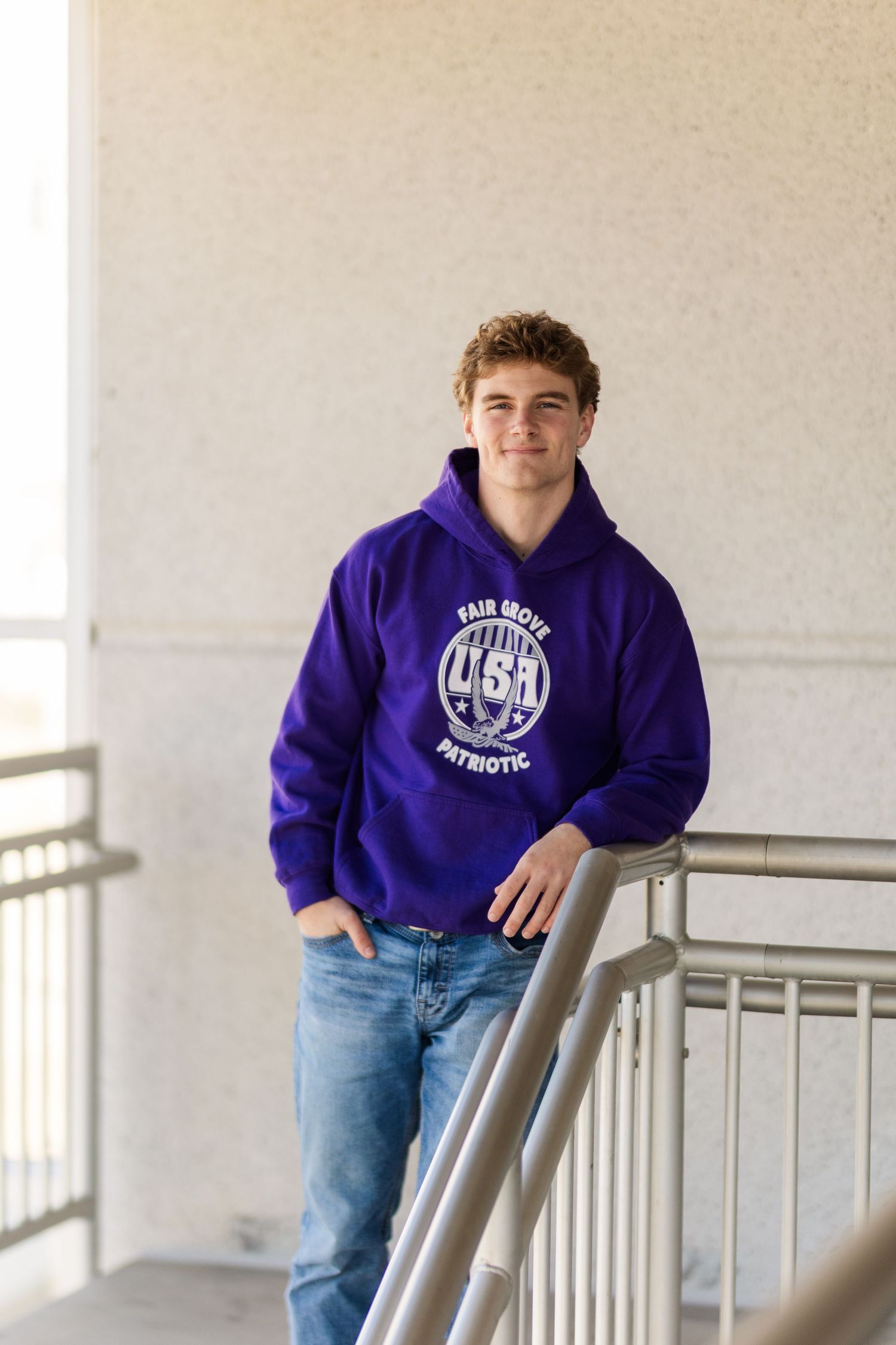 A person wearing a purple university hoodie and jeans leans against a railing in a bright hallway.