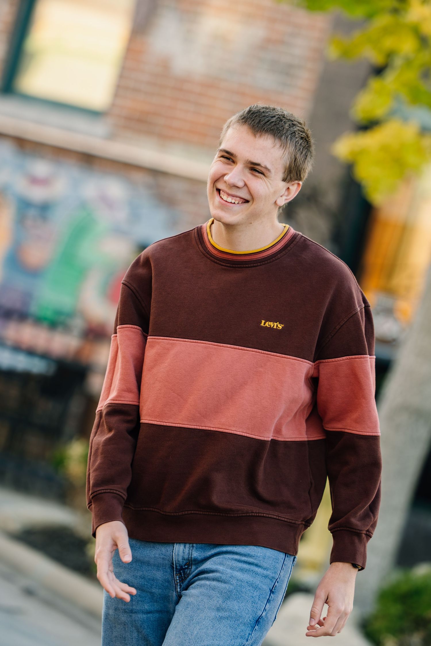 A person wearing a brown and burgundy color-block sweatshirt with blue jeans stands outdoors in natural lighting.