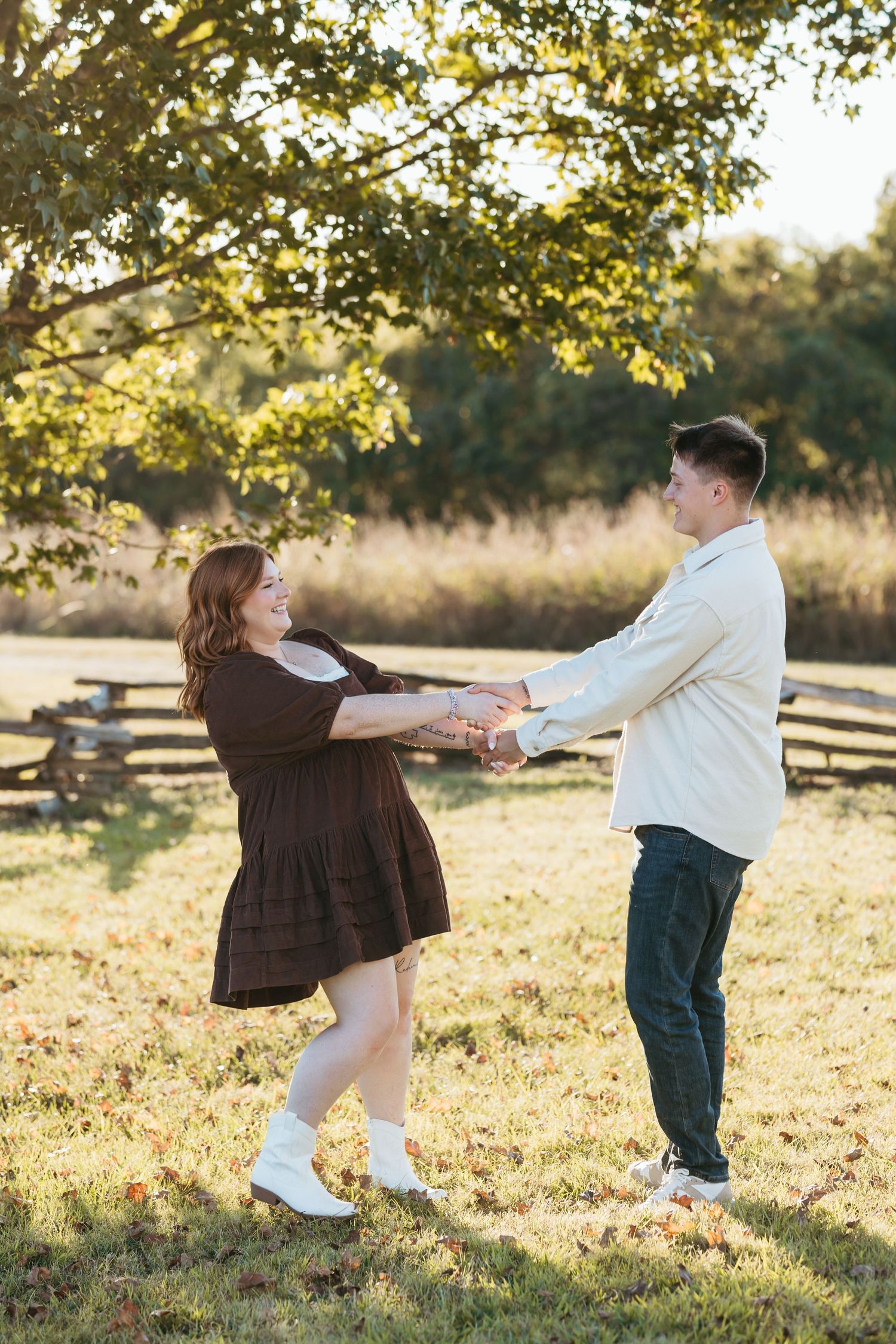 Couple dancing and spinning in sunlit field with wooden fence and autumn trees behind them.