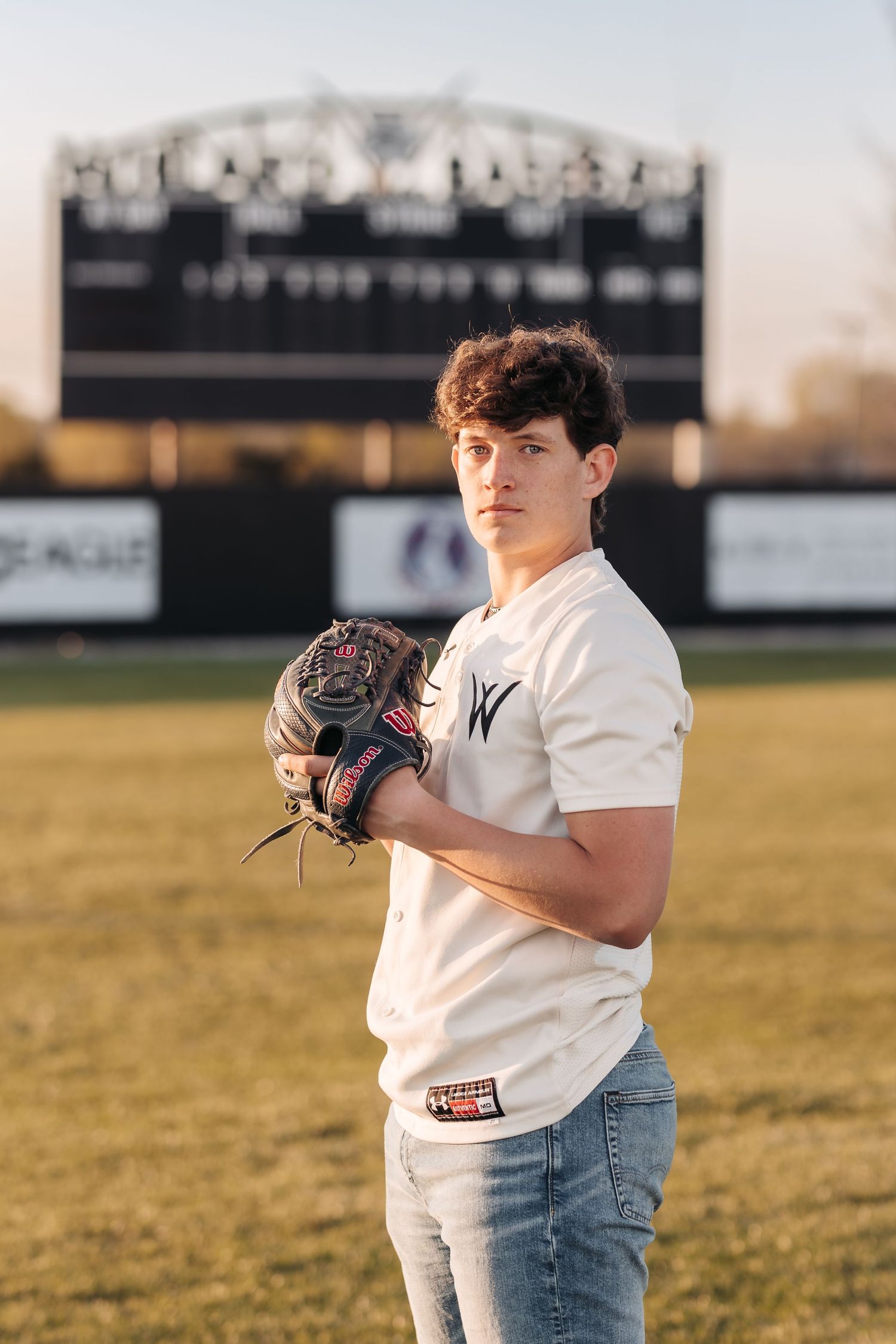 Baseball player in white jersey poses with glove on field at sunset with scoreboard behind.