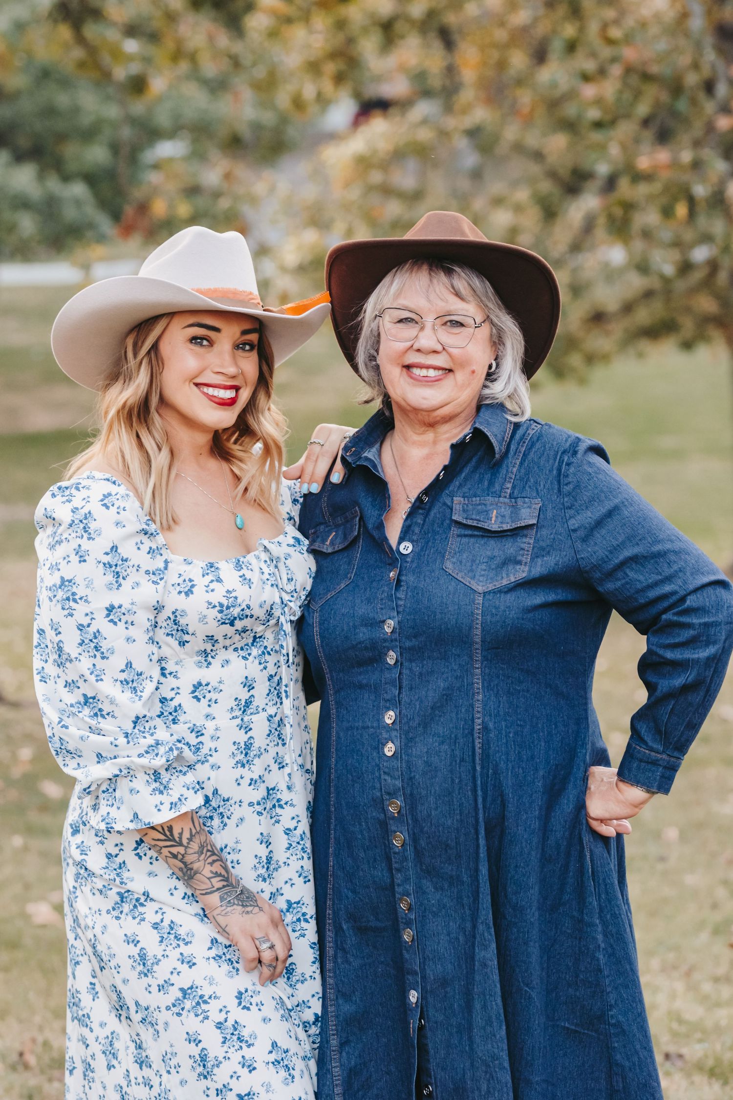 Two people pose together outdoors in western style, wearing a denim dress and floral print dress with cowboy hats.