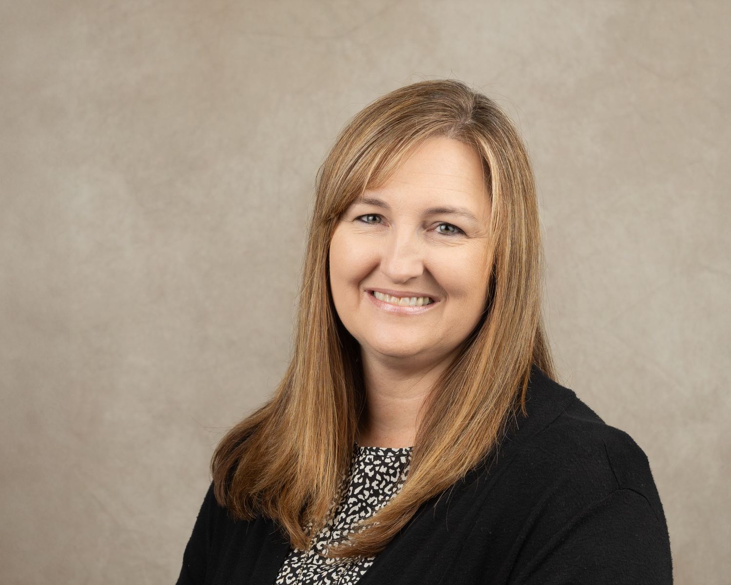 Professional headshot of a person wearing a patterned top and black blazer against beige background.