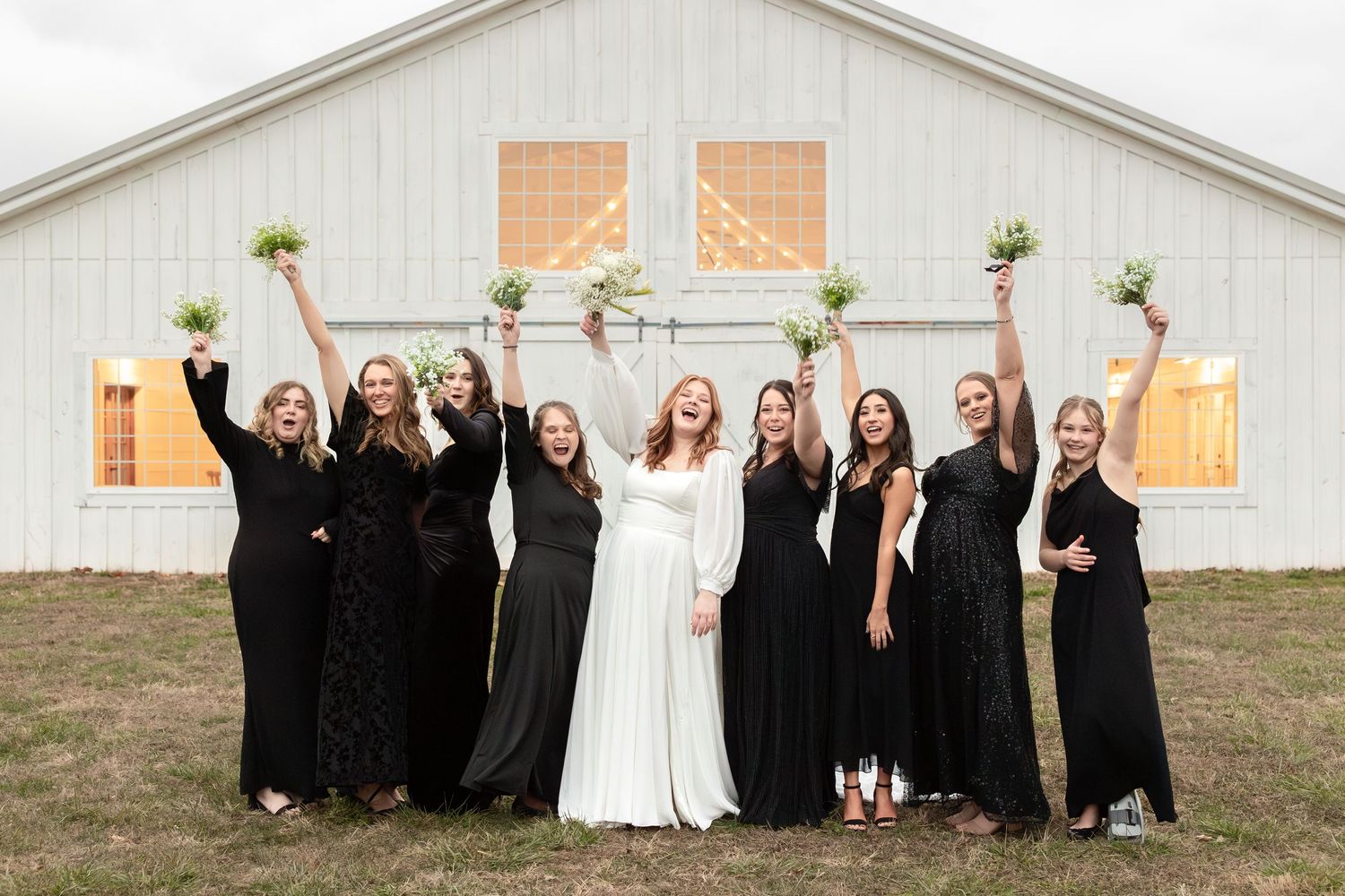 Bridal party in black dresses celebrates with raised bouquets in front of a white barn.
