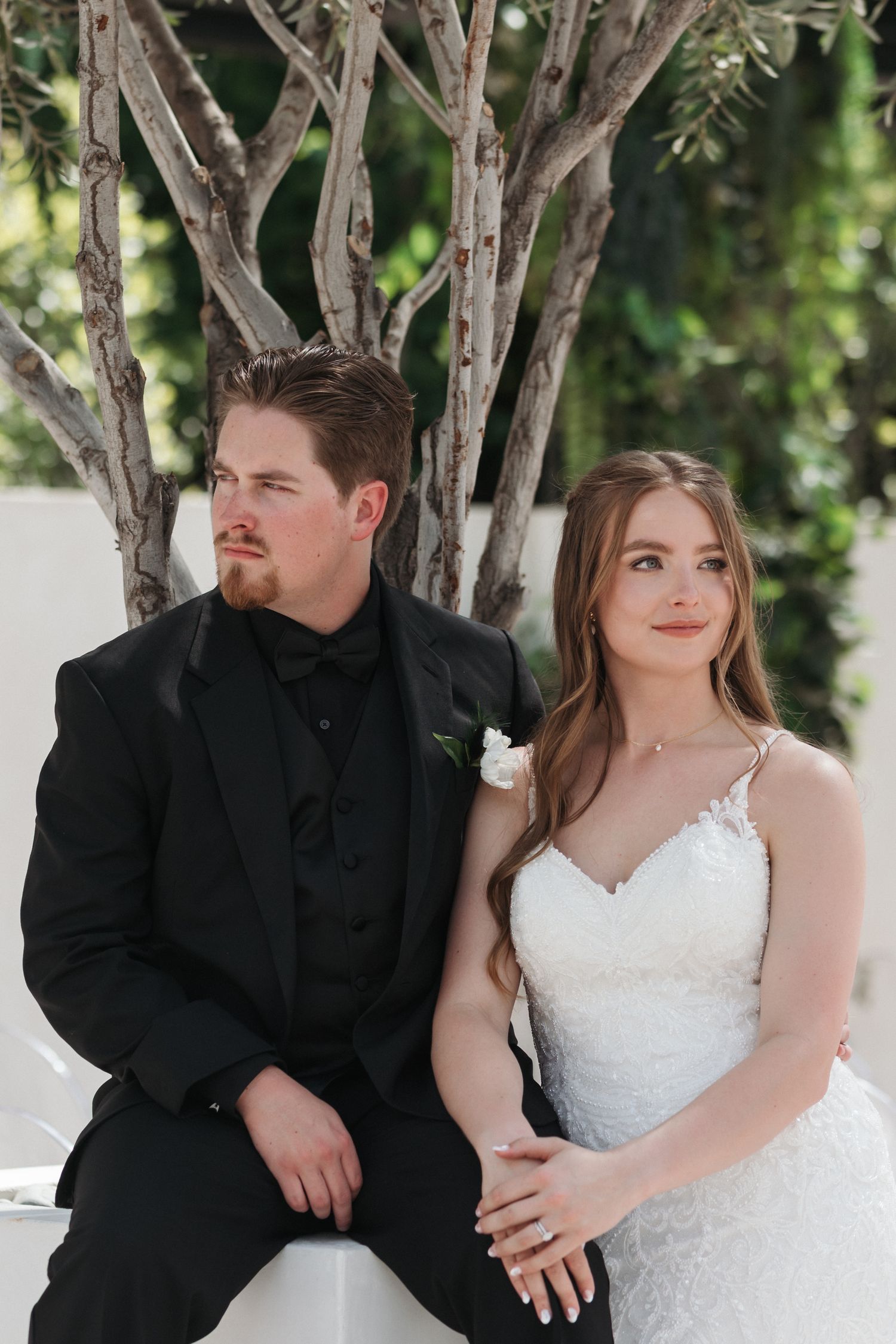 Elegant wedding portrait of couple sitting together against white wall with rustic branches.