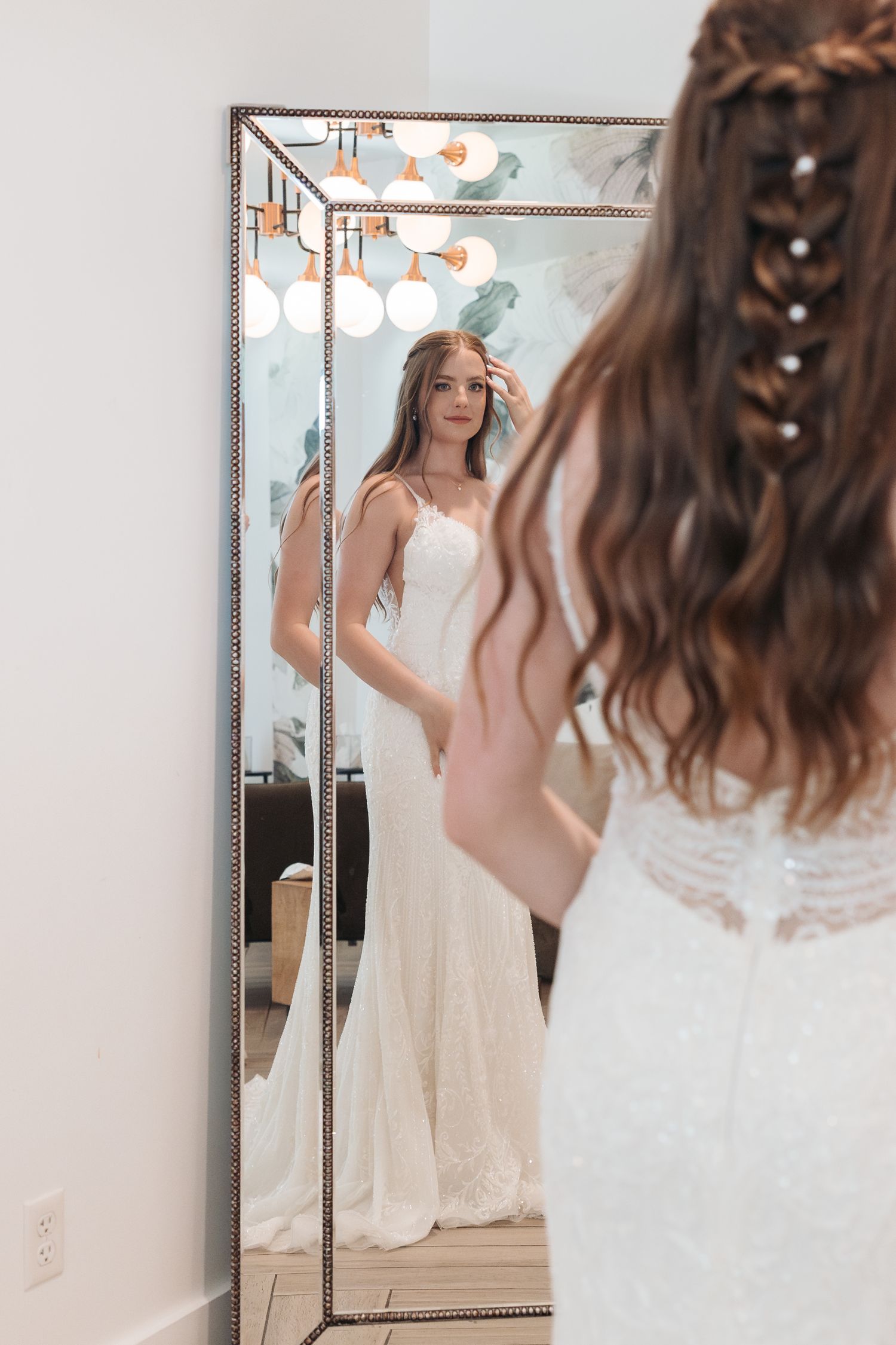 Reflection of bride in elegant white wedding gown looking at herself in a full-length mirror.