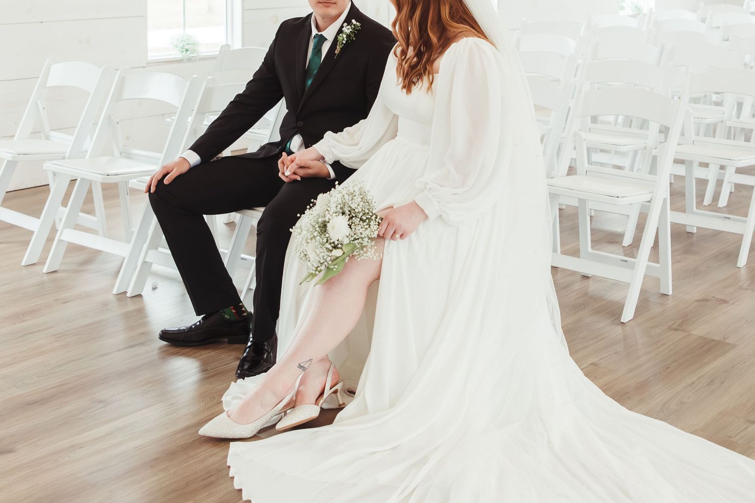 Bride and groom sit together showing their shoes at a wedding ceremony with white folding chairs.
