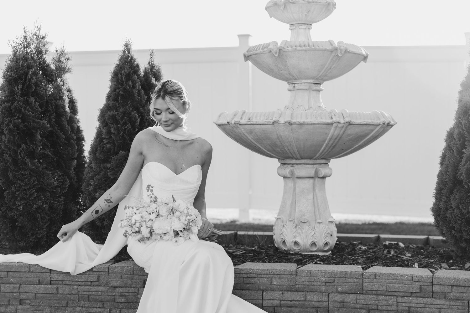 Elegant bride in strapless gown holding bouquet beside ornate stone fountain in black and white.