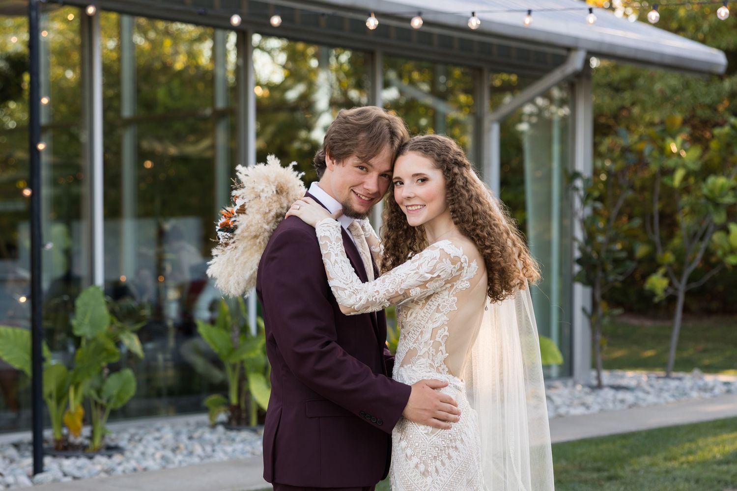 Bride and groom embracing outdoors at elegant wedding venue with string lights and lush greenery backdrop.