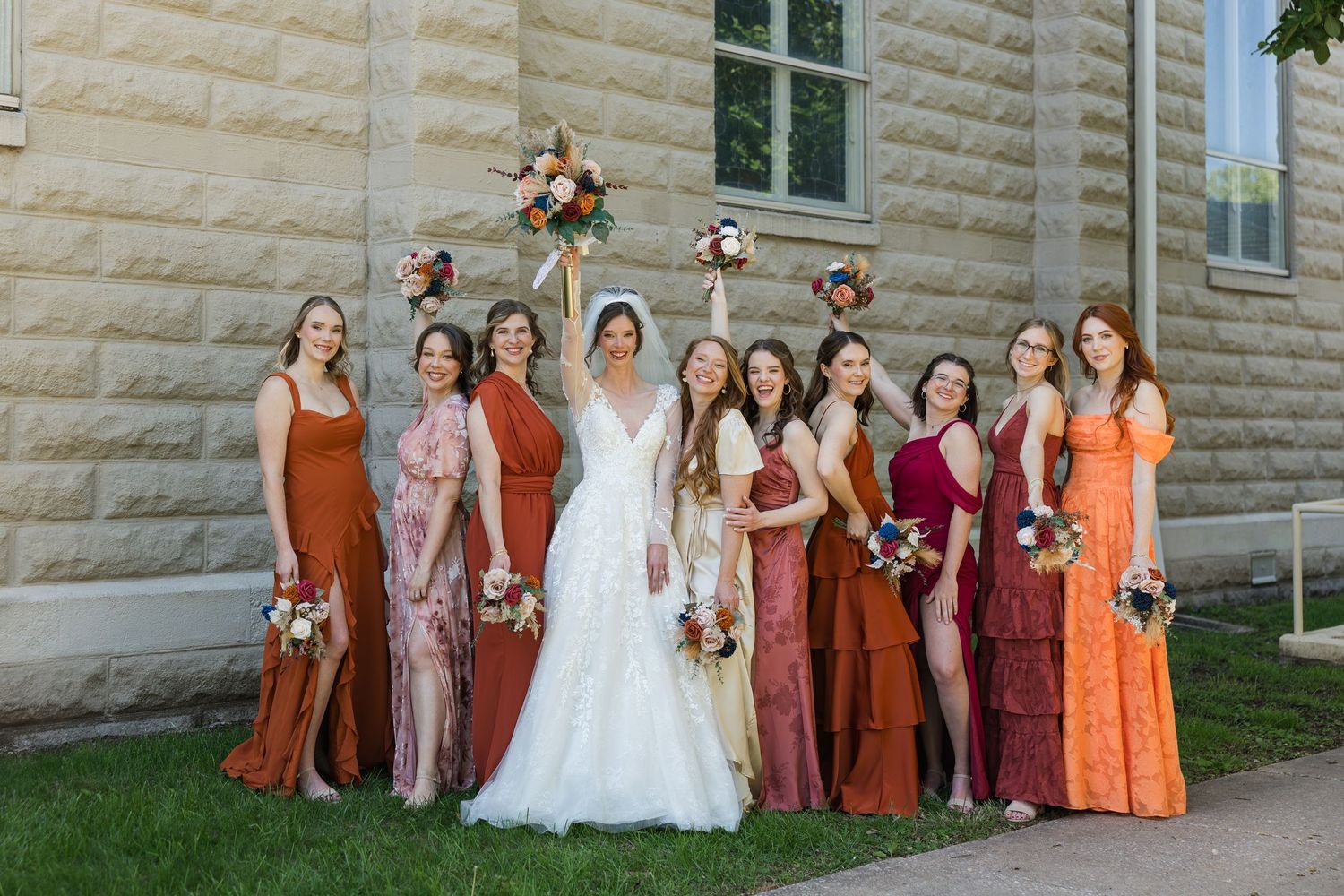 Bride in white gown poses with bridesmaids in mismatched rust and orange dresses outside a stone building.