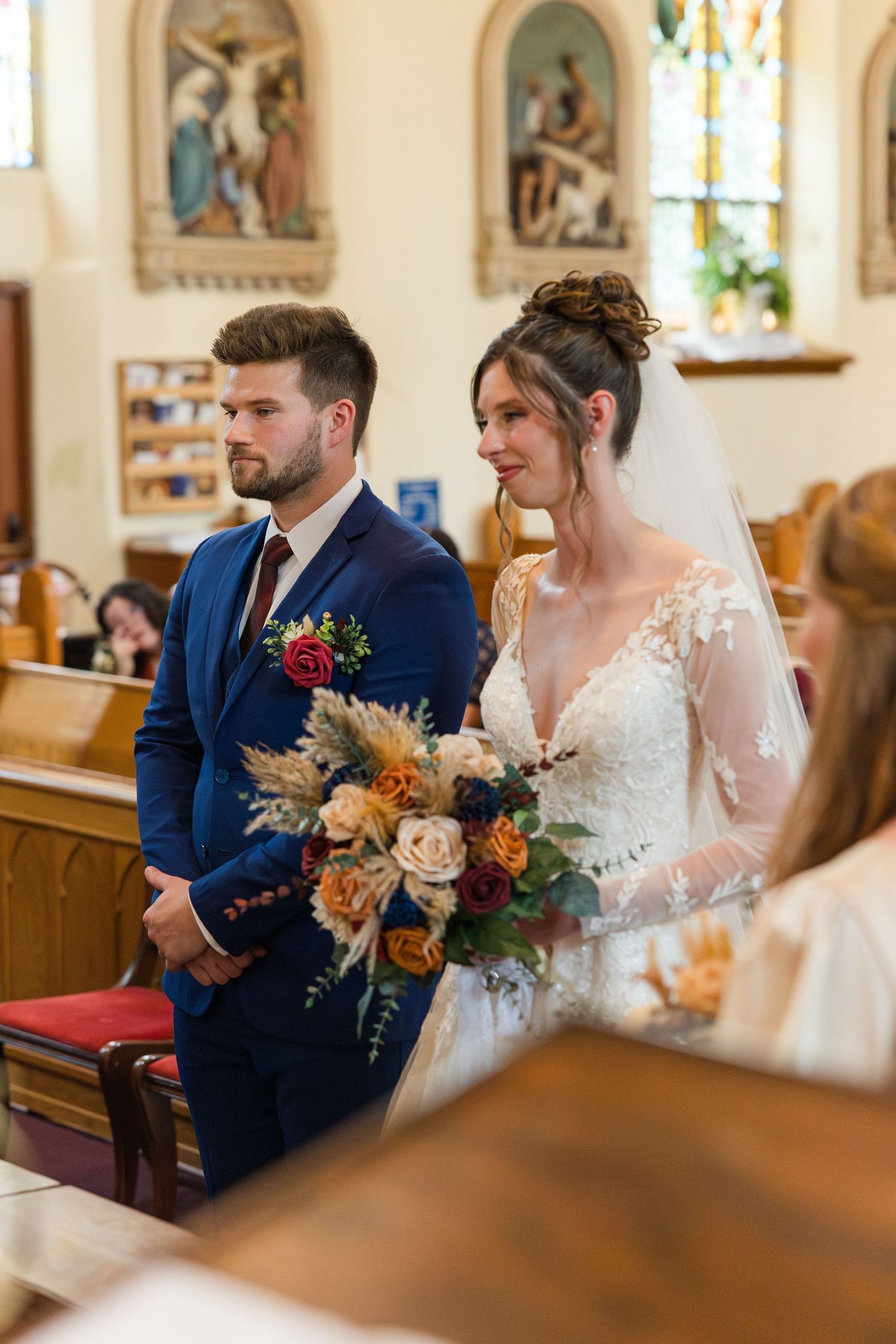 Bride and groom stand at church altar, she holds autumn bouquet in lace gown, he wears navy suit.